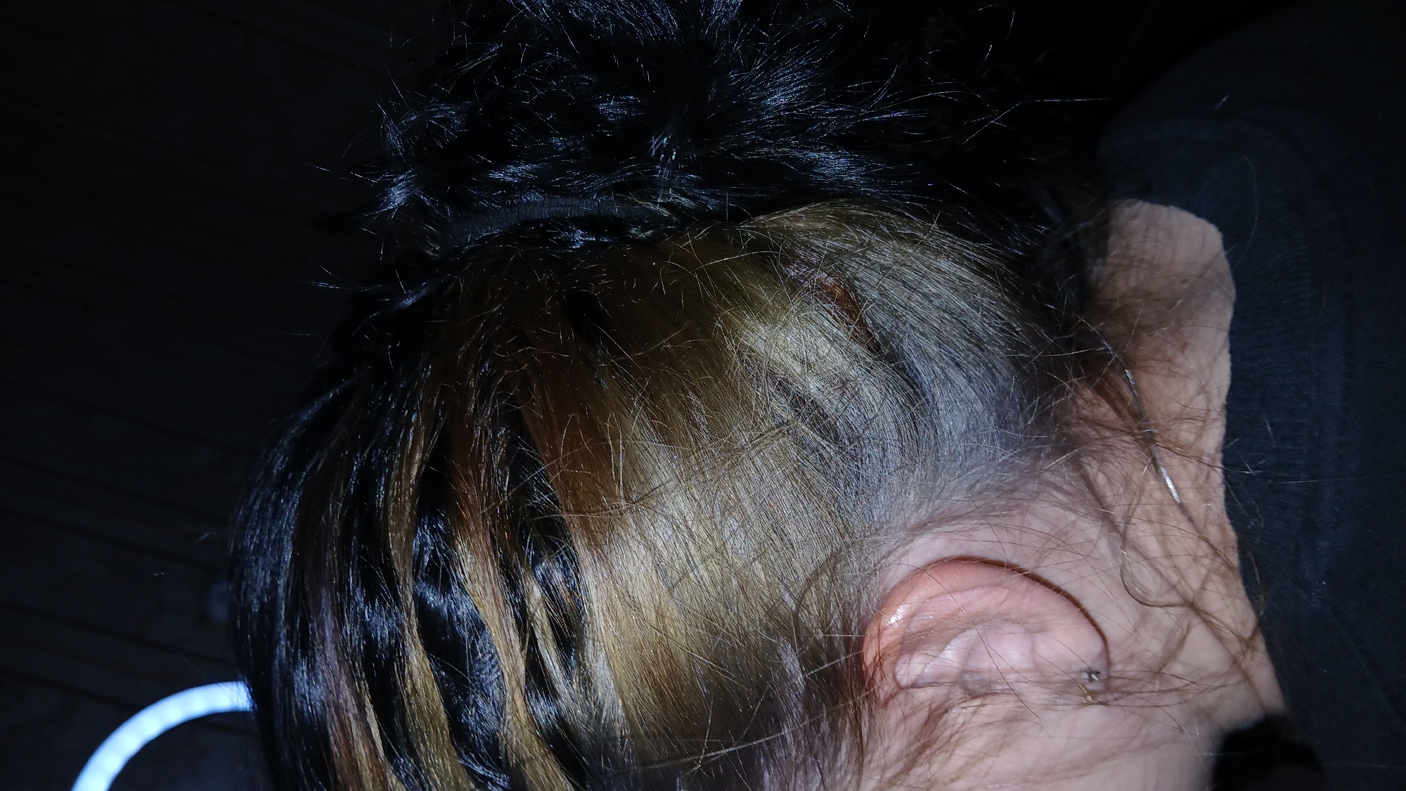 A close-up shot focuses on the back of a person's head and their right ear. The hair is styled in a high bun, with black hair on top and lighter brown and blonde highlights underneath. The hair is held in place with a black hair tie. The person's ear is visible with a small piercing. A dark garment, possibly a hoodie, is partially visible behind the head. The background is dark and somewhat blurred, with a hint of an illuminated ring-shaped object in the lower left corner. The location is unknown. There is no indication this image was taken in Dnipro, Ukraine.