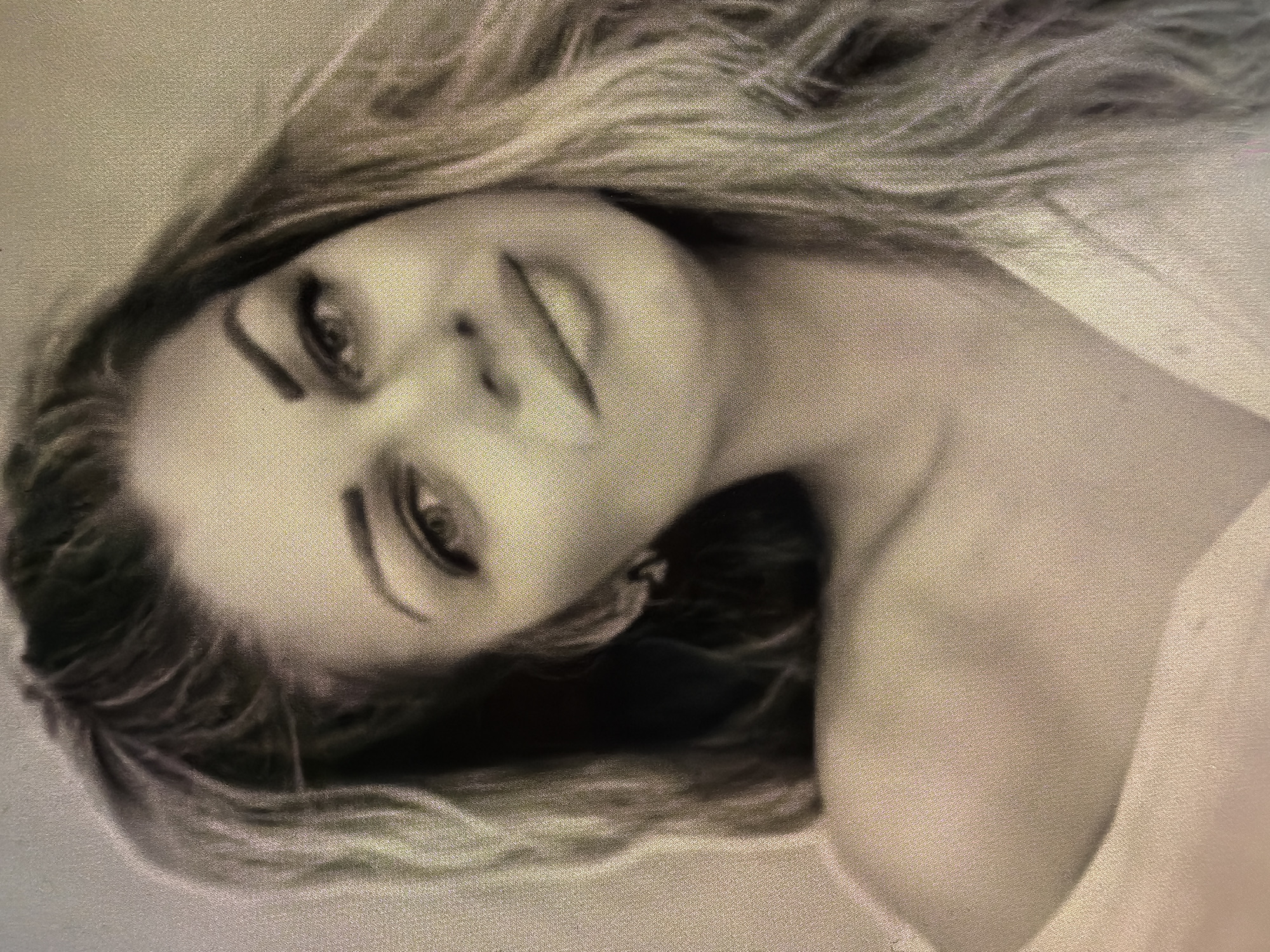 The image is a black and white portrait of a woman. She has long, flowing hair and is wearing what appears to be a simple dress or top. Her head is tilted slightly, and she is looking directly at the camera. The background is plain and out of focus. The location of the image is not discernible from the content.