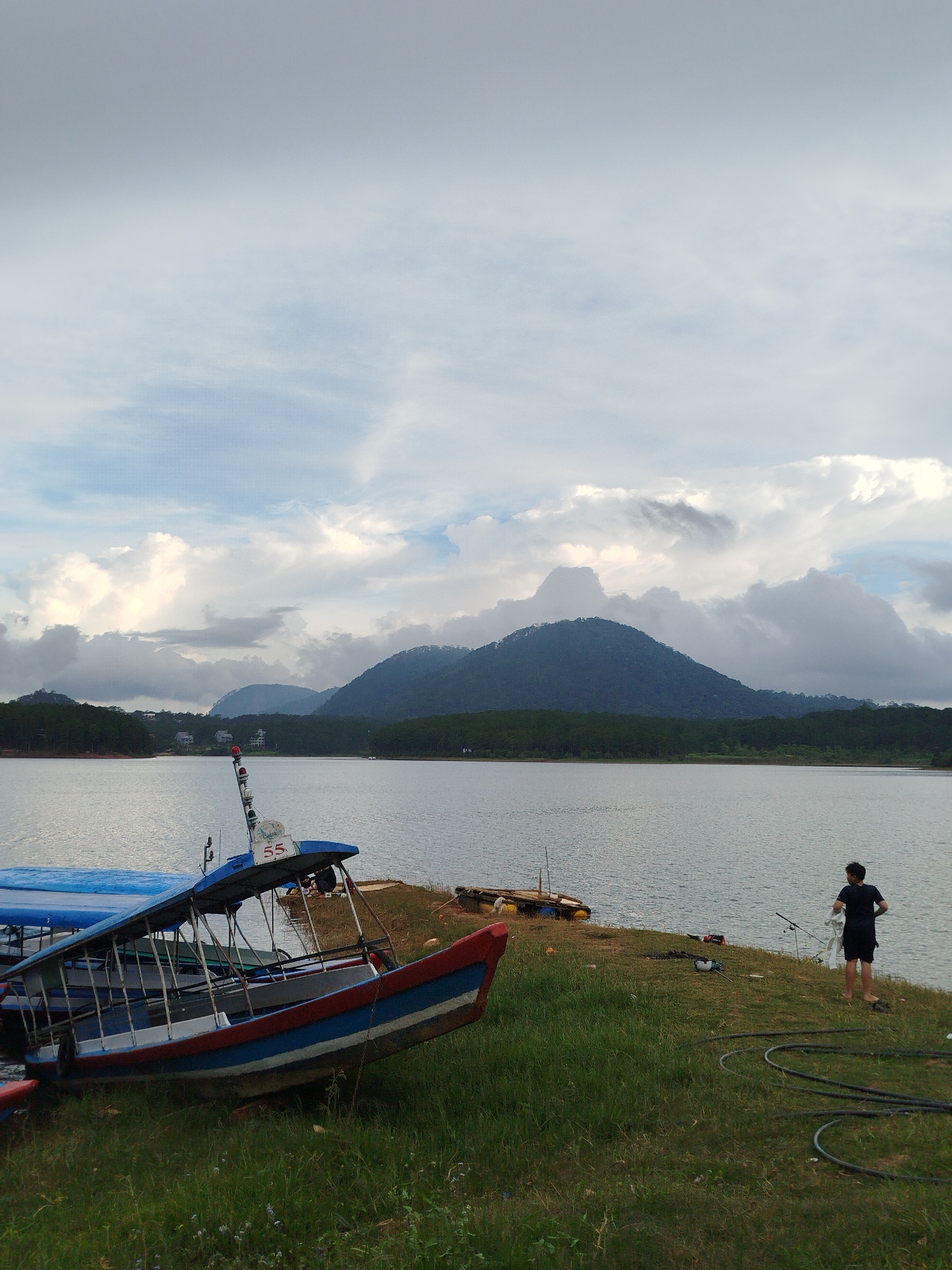 The image depicts a serene lake scene in Da Lat, Vietnam, likely Tuyen Lam Lake, known for its scenic beauty and recreational activities. In the foreground, a rowboat with a blue and red hull rests on a grassy embankment, its canopy providing shade. The boat is positioned at an angle, leaning towards the water. Further back, another blue-covered boat is partially visible.
The lake stretches out to a distant shoreline lined with lush green trees and rolling hills covered in dense vegetation. Several mountains, their peaks shrouded in a soft, hazy light, rise majestically in the background. The sky above is dramatic, filled with large, cumulus clouds tinged with shades of gray and white, suggesting an overcast or approaching storm.
On the grassy bank near the water's edge, a lone individual, dressed in a black t-shirt and shorts, is engaged in an activity that appears to be related to fishing or boat maintenance, holding a white cloth. Several black hoses are coiled on the grass nearby, and a fishing rod is set up. The overall atmosphere is tranquil and natural, capturing the essence of Da Lat's picturesque landscapes.