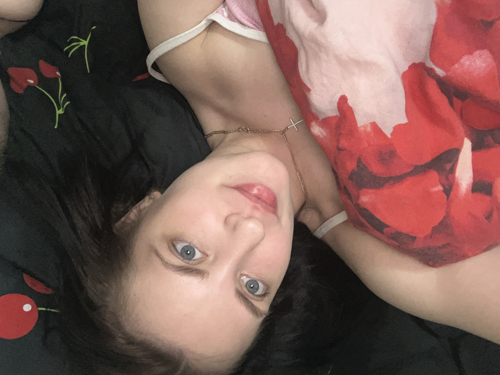 A high-angle, close-up photograph captures a woman lying down, looking directly at the camera. She has light skin, striking blue eyes, and dark hair, part of which is visible around her head. Her lips have a soft pink hue. She wears a delicate gold-toned chain necklace with a small cross pendant, which rests on her collarbone.
She is dressed in a light pink top with white trim. A larger piece of fabric with a vibrant red and white abstract or floral pattern is visible, possibly a blanket, duvet, or another piece of clothing covering part of her body. She is positioned against a dark fabric, likely a bedsheet or blanket, which features stylized red floral or cherry-like designs with green stems.
The scene appears to be indoors, suggesting a personal and relaxed setting, possibly a bedroom. The woman's expression is calm and neutral. There are no discernible elements in the image to indicate the time of day or weather conditions. No text is visible within the image. The location context provided is Markovskiy, Russia.