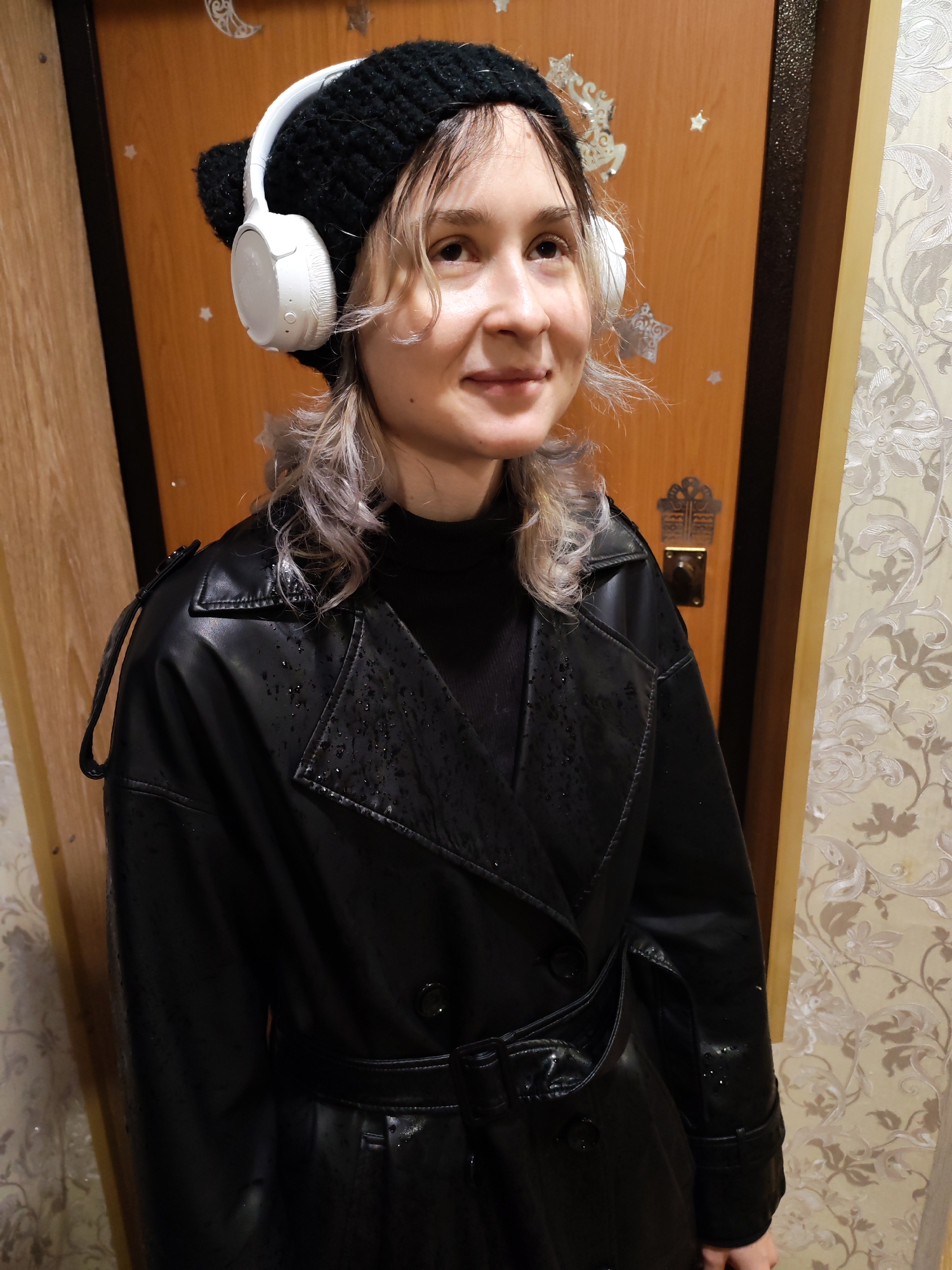 A young woman is depicted in a medium shot, standing indoors, likely in an entryway or hallway of a residence in Chaikovsky, Russia. She has fair skin, wavy, light-colored hair, and is looking towards the viewer's left with a gentle, pleasant smile.
She is wearing a black knitted beanie, white over-ear headphones, a black high-neck top, and a black faux leather trench coat. The trench coat features a double-breasted front, shoulder epaulets, and a belted waist. Small water droplets are clearly visible on the surface of the coat, particularly on the shoulders and arms, indicating it is wet, possibly from rain.
The background features a light brown wooden door adorned with several reflective silver star and crescent moon stickers. A decorative metal escutcheon surrounds a doorknob on the door. To the right, a wall is covered in patterned wallpaper with a subtle floral or vine design in light beige and white tones. A cream-colored door frame separates the door from the wallpaper. The setting suggests a casual, domestic moment, perhaps after coming inside on a damp day. No visible text is present in the image.