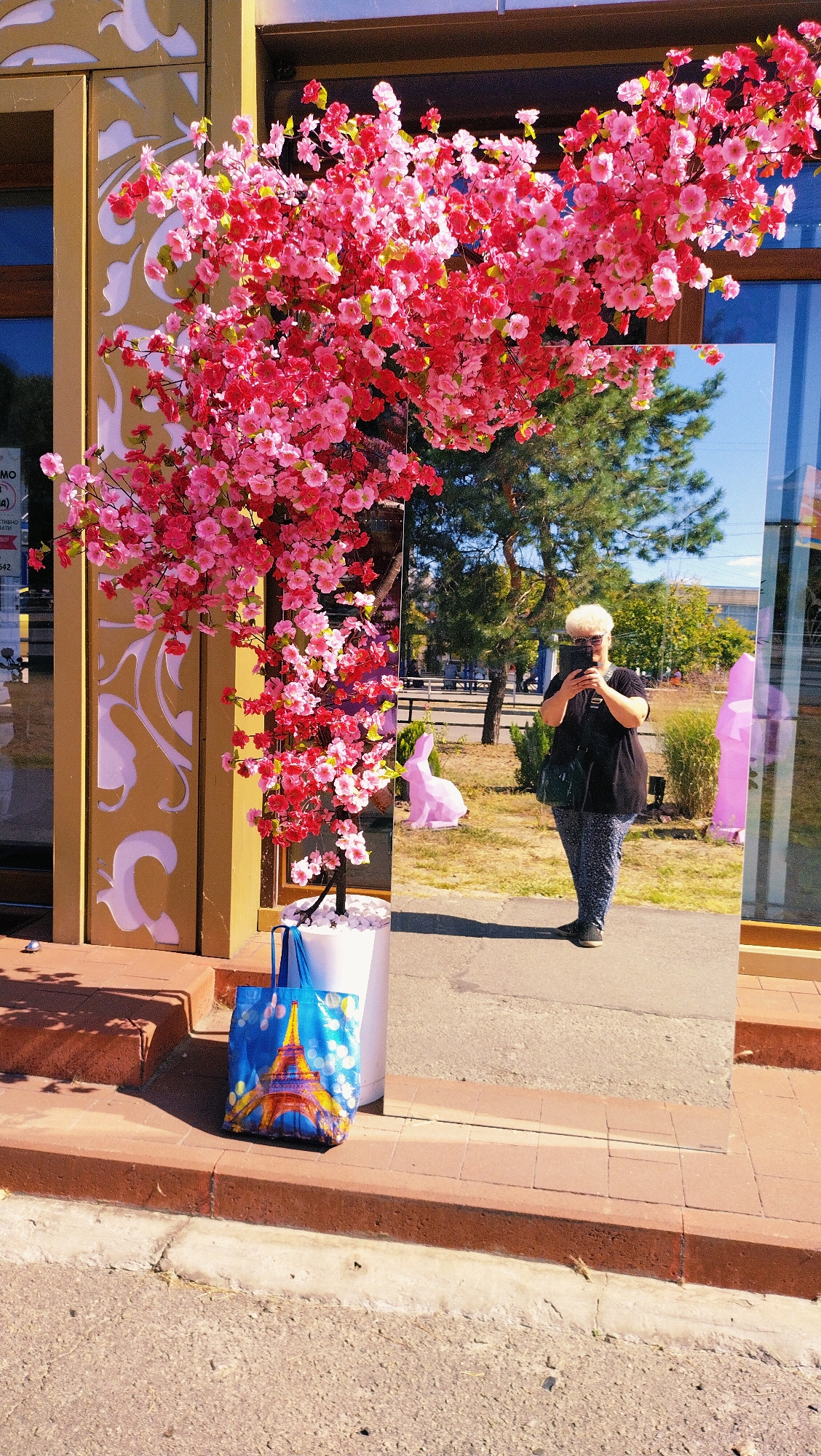 A person with white, curly hair wearing sunglasses, a black t-shirt, and patterned pants is standing in front of a large mirror, taking a selfie with a dark-colored mobile phone. The mirror reflects the person, a pink rabbit statue, trees, and a building in the background. In the foreground, to the left of the mirror, is a white planter with a faux cherry blossom tree. Beside the planter is a blue tote bag with an image of the Eiffel Tower on it. The scene appears to be on a sidewalk in front of a building with decorative elements on the exterior wall, possibly in the city of Horishni Plavni, Ukraine, given the context. The ground is paved with asphalt and brick.