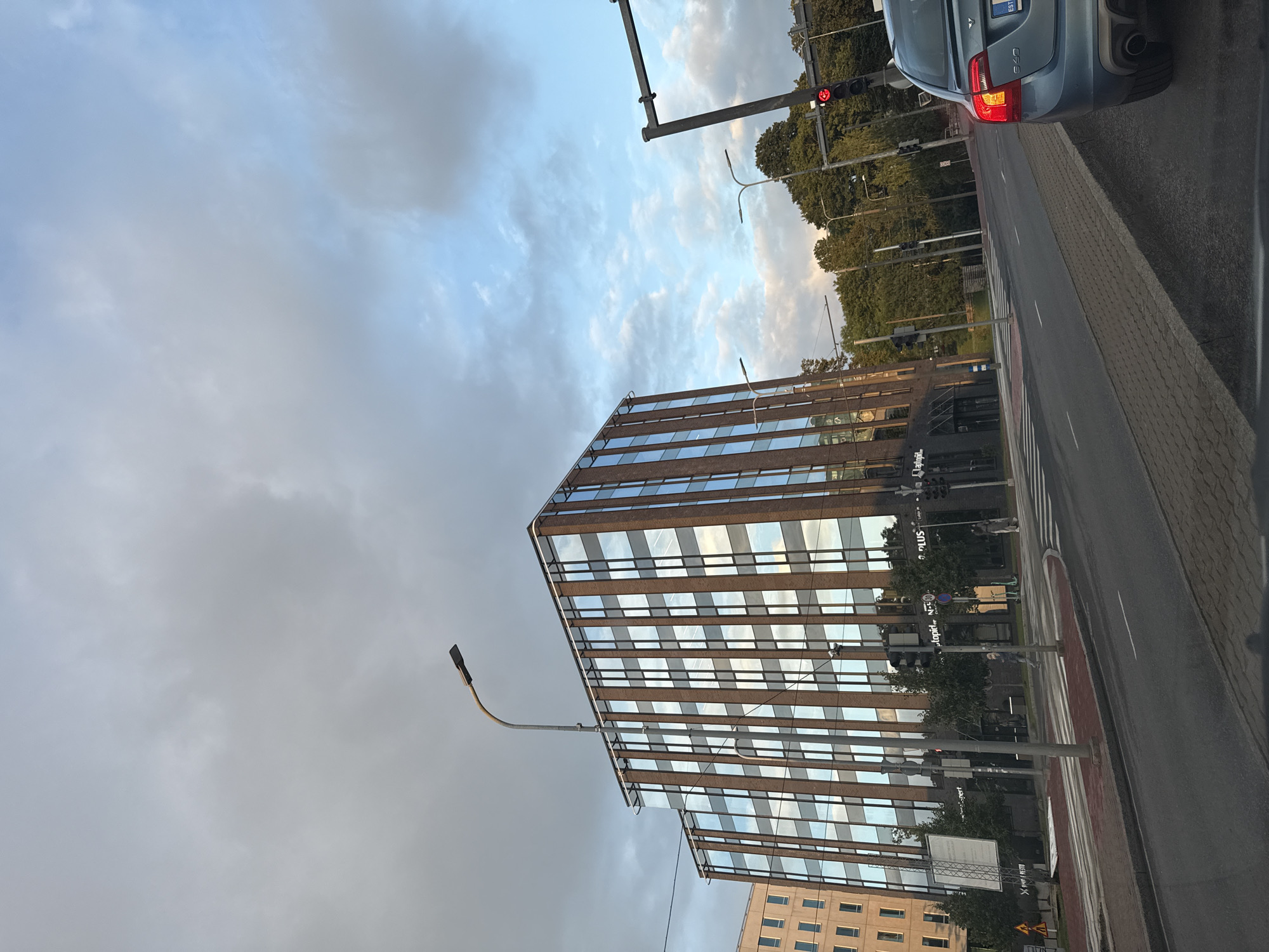 The image shows a view of a street in Tallinn, Estonia. In the foreground, a gray road with a brick sidewalk is visible. To the right, the rear of a blue Volvo S60 is parked, with its brake lights illuminated. In the mid-ground, a modern, multi-story building with a glass facade and vertical brown accents stands prominently. Across the street, there is a pedestrian crossing. Further down the street, trees line the road, and a traffic light with a red signal is visible. The sky above is partly cloudy. The building has signage that is partially visible, including "autoopils", "PLUS", and "Lutsugi".