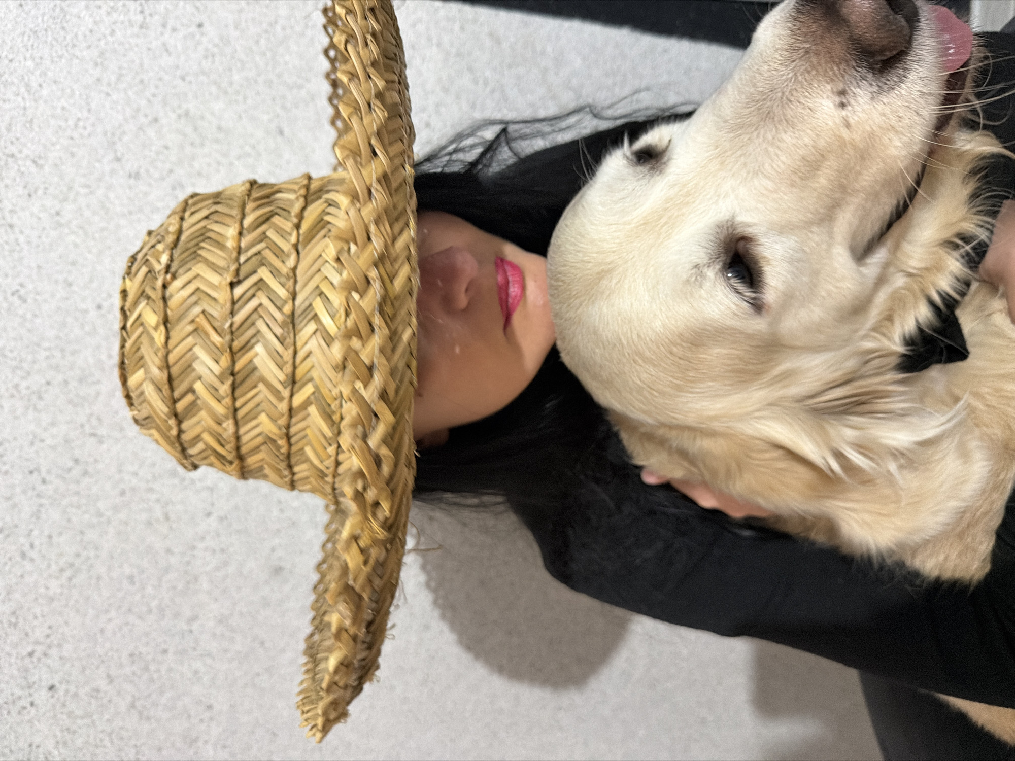 A woman and a light-colored Golden Retriever dog share an intimate, close-up moment in this image. The woman is positioned on the left side of the frame, viewed from a high angle, possibly lying down or leaning back. She has long dark hair and is wearing a black top. A woven straw hat is prominently featured on her head, its wide brim extending towards the left and partially obscuring her face. Her lips are visible beneath the hat's brim, accentuated by vibrant pink lipstick.
The Golden Retriever is pressed closely against the woman's face, its head near her temple and cheek, suggesting a tender interaction. One of the dog's eyes is clearly visible, conveying a gentle expression. Its pink tongue is slightly exposed, hinting at a lick or an affectionate gesture towards the woman. A hand, likely the woman's, is seen gently supporting the dog's head or body.
The background consists of a plain, light-colored wall with a subtle textured appearance, indicating an indoor setting. The lighting is soft and even. The overall scene conveys a feeling of affection, warmth, and strong companionship between the woman and her pet. No visible text is present in the image.