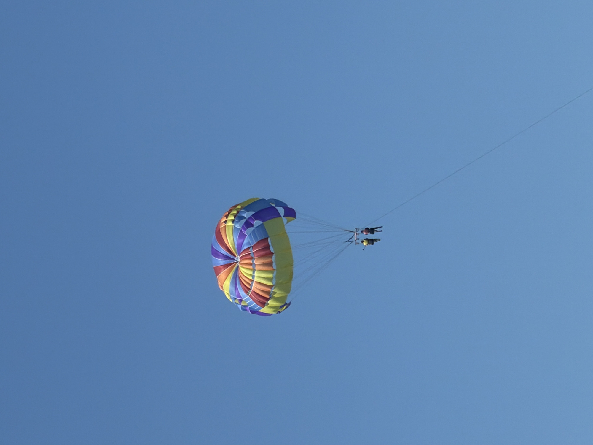 A colorful parachute with sections of yellow, orange, red, purple, and blue is soaring against a clear, bright blue sky. Two people, appearing as dark silhouettes, are attached to the parachute and are parasailing. The parachute is positioned in the lower left portion of the frame, angled upwards and to the right, suggesting it is high in the sky. A thin tow rope extends diagonally across the upper right portion of the image, connecting the parachute to an unseen boat or vehicle. The overall scene depicts a sunny day, likely in the afternoon, with excellent visibility. The clear sky and the activity of parasailing suggest a recreational setting, perhaps a beach resort or a coastal area. No text is visible in the image. The image evokes a sense of fun and adventure.