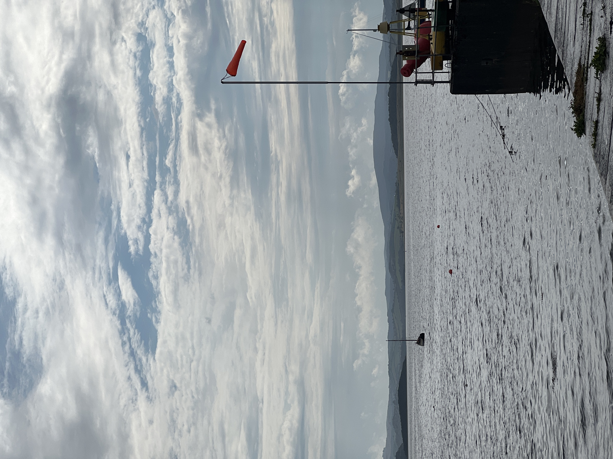 The image depicts a cloudy sky over a body of water. In the foreground, a dock or pier is visible on the right side, with a tall pole topped with an orange windsock. The water is choppy, and a single sailboat is moored in the distance. Hills and landforms are visible on the horizon. The scene appears to be a coastal or harbor area. The specific location within Terhillion, Ireland cannot be determined from the image alone.