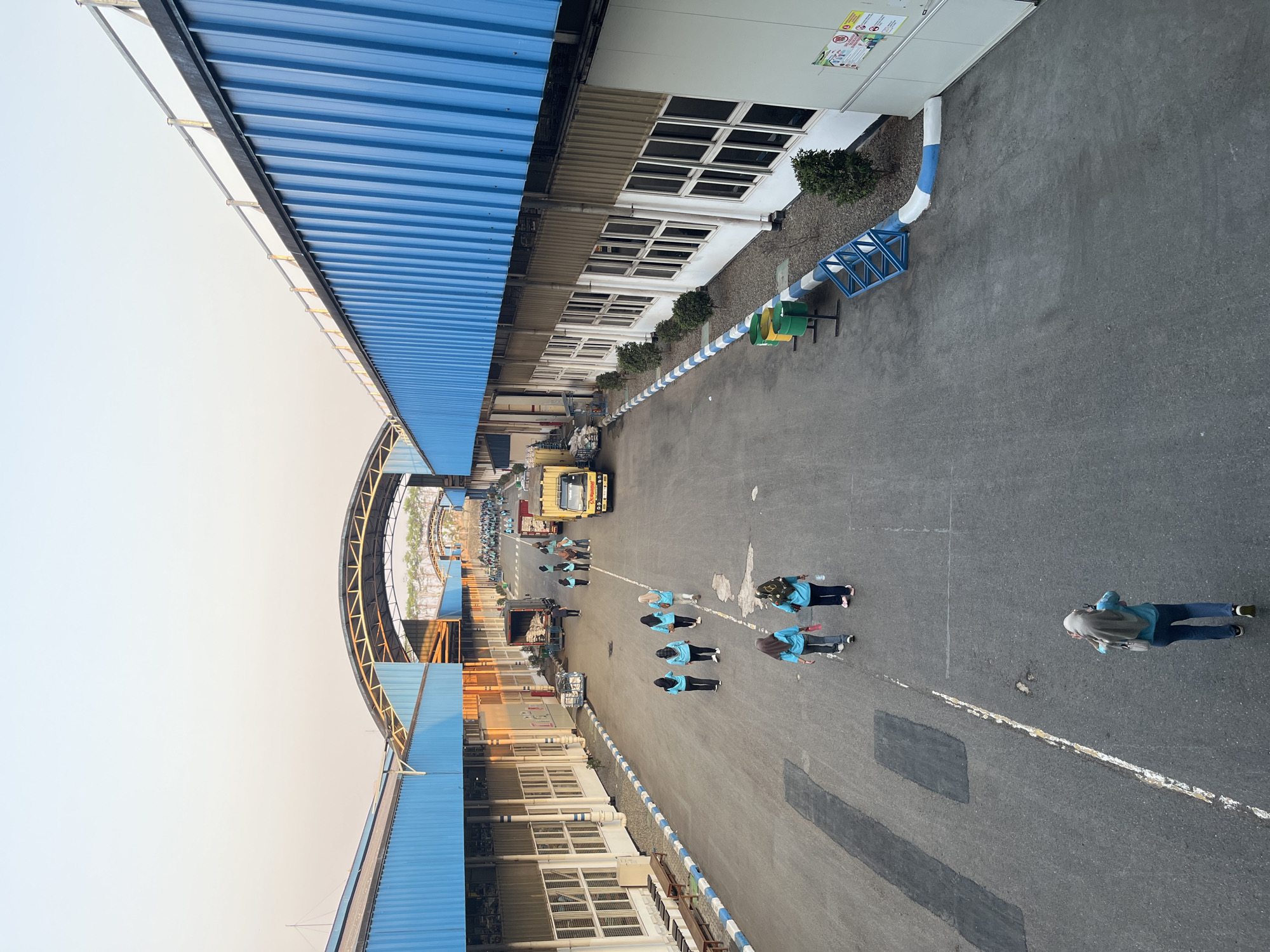 The image depicts an outdoor area within an industrial facility, likely located in Pelang, Indonesia, given the context. The scene is dominated by long, low-rise buildings with prominent blue corrugated metal roofs and white walls, interspersed with open paved areas. Several people are walking in various directions across the paved expanse. Most of them are wearing blue shirts, suggesting they are likely employees or visitors to the facility. Some individuals are walking individually, while others are in small groups.
In the background, a yellow truck is parked, and another truck, possibly a delivery vehicle, is visible further down the road. The architecture features arched roof structures spanning between buildings. The ground is asphalt, with painted white lines indicating a pathway or road. The overall atmosphere suggests a working environment, possibly during the late afternoon given the warm lighting and long shadows. The presence of trash bins and some landscaping elements like small trees and bushes indicate that the area is maintained. The sky is clear and light blue, transitioning to a warmer hue near the horizon, indicative of either sunrise or sunset.