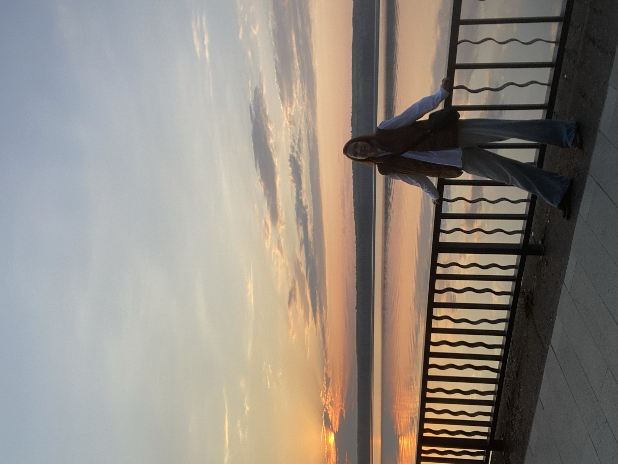 A woman stands by a railing with a sunset and lake in the background. The woman is wearing a white shirt, a brown vest, and light-colored pants. She is leaning on the railing, looking out at the sunset. The railing is black metal with a wavy design. The lake is calm, with a reflection of the sunset on its surface. The sky is a mix of colors, including orange, pink, and blue. The picture was taken in Chaikovsky, Russia.