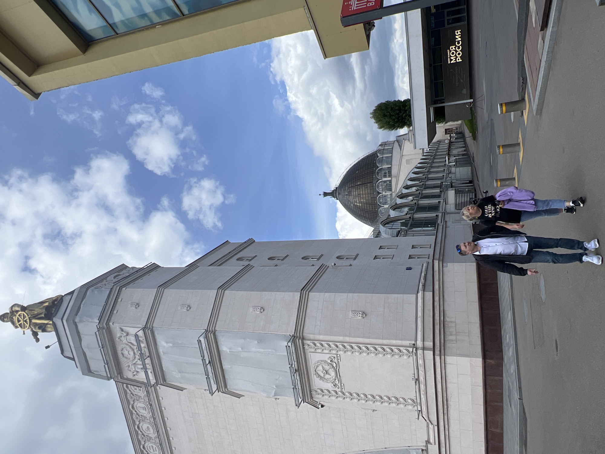 The image shows a low-angle view of the Pavilion No. 1 "Central" at VDNKh, Moscow, Russia. The pavilion is a tall, rectangular building with a tower topped by a golden statue. Adjacent to the pavilion is a domed building, presumably another structure within VDNKh. Two individuals are walking on the sidewalk beside the pavilion, a woman with a purple jacket and a man in a jacket and white shirt. The sky is visible, with scattered clouds. A building with signage "моя Россия" ("My Russia") is also partially visible.