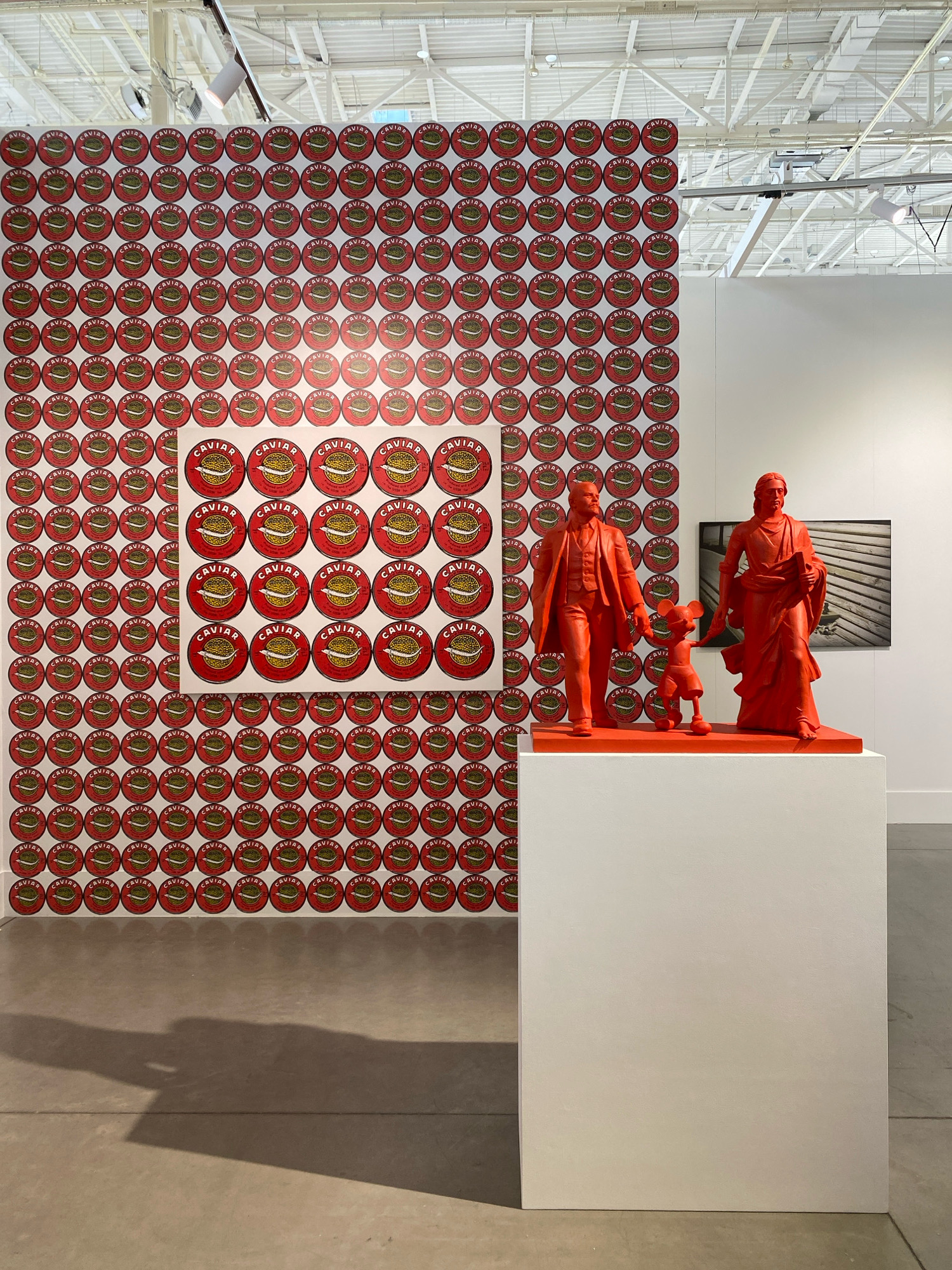 The image displays an art exhibition inside a building with a high ceiling featuring industrial lighting. On the left, a large wall is covered in a repeating pattern of circular red labels, each bearing the word "CAVIAR" and an image of a fish. A framed artwork, a grid of similar caviar labels, is mounted on this patterned wall. To the right, on a white pedestal, are three bright orange sculptures: a man in a suit, a dog, and a woman in a flowing gown. In the background, a framed photograph of wooden planks is visible on a white wall. The floor is concrete, and a shadow is cast across it from the left side of the frame. The scene is set in the city of Khimki, Russia.
