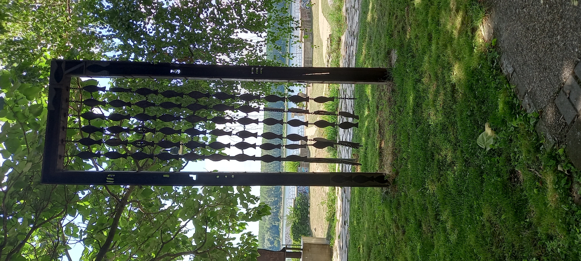 The image captures a metal sculpture outdoors, likely in the city of Silistra, Bulgaria, given its riverside setting. The sculpture, composed of dark metal, features two vertical posts connected by a horizontal beam at the top, forming a rectangular frame. Hanging from the horizontal beam are multiple chains, each adorned with a series of stylized fish-shaped metal cutouts. These fish are arranged in rows, creating a decorative screen.
The sculpture is situated in a grassy area with a paved pathway running along the bottom edge of the frame. In the background, a body of water, presumably the Danube River, is visible. Trees with lush green foliage overhang the sculpture, casting dappled shadows on the ground. Beyond the river, a tree-lined bank is discernible, with some buildings or structures present on the far side. The sky is clear and blue.
There are no people or animals visible in the image. The focus is on the sculptural element and its immediate surroundings. The overall impression is one of an outdoor public art installation in a natural, waterside environment.