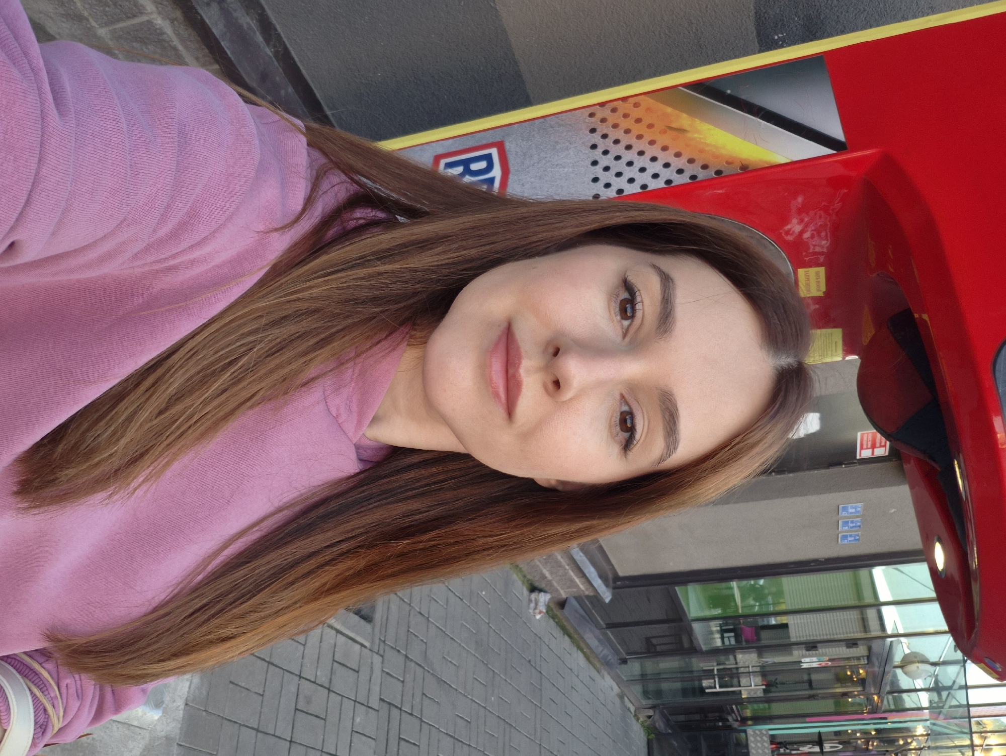 A medium shot of a woman with long, brown hair, wearing a pink hoodie, taken from the chest up. She is standing outdoors, in front of a red and yellow structure, possibly a vending machine, with a gray brick wall and glass-fronted building behind her. The ground is paved with gray rectangular tiles. The woman's head is turned slightly to her left, and she is looking towards the camera. The scene appears to be a street in the city of Kudrovo, Russia.