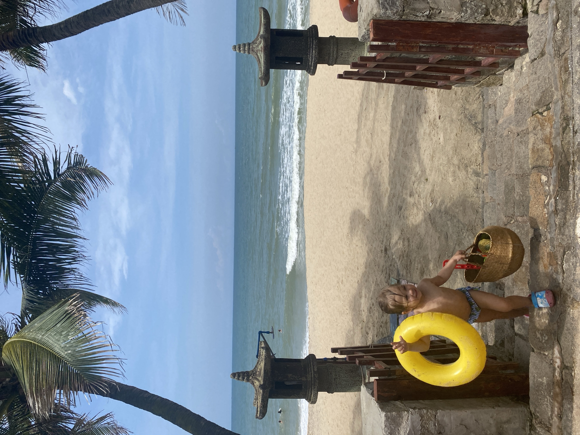 A young child is walking up stone stairs towards a sandy beach, holding a yellow inflatable ring in their left hand and a woven basket in their right. The child is wearing striped swim trunks and colorful sandals. In the background, the ocean waves are breaking on the shore, and palm trees frame the scene under a partly cloudy blue sky. To the left and right of the stairs, there are stone lanterns. The location appears to be a beach in Phu Thuy Ward, Vietnam.