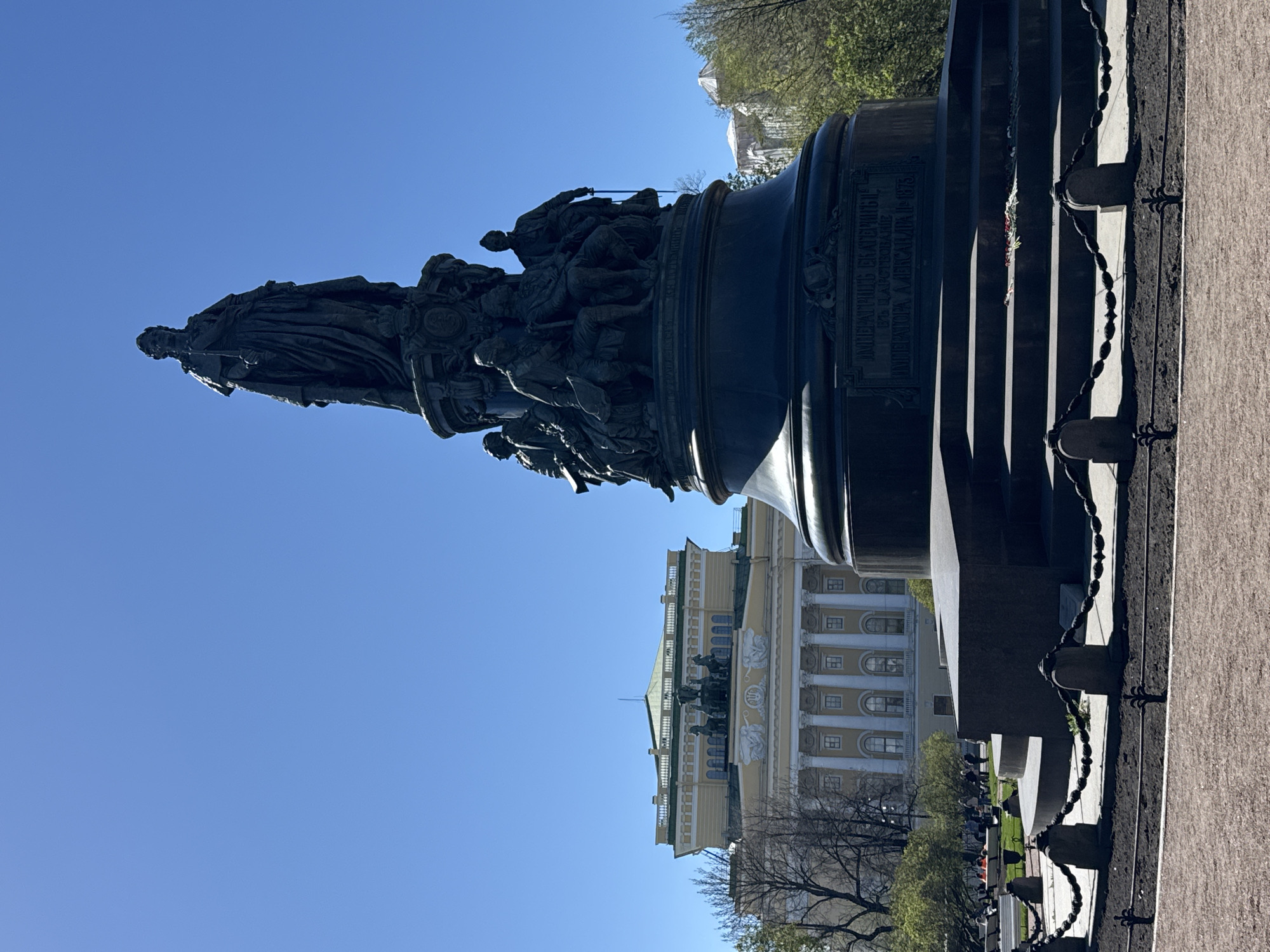 The image shows the Catherine the Great Monument in Saint Petersburg, Russia, set against a clear blue sky. The statue of Catherine the Great stands atop a large, elaborately decorated pedestal with multiple figures. The facade of a building with a light-colored exterior and architectural details is visible in the background. A fence made of thick metal chains is placed around the monument. People are walking. A paved area is visible in the foreground.
