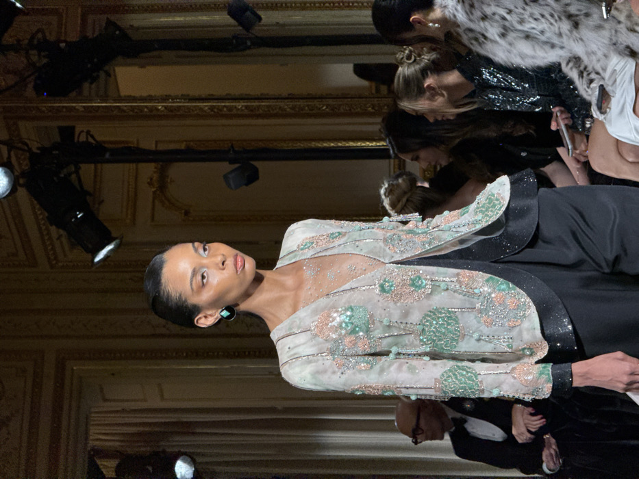 A model poses on a runway, her elegant attire featuring a sheer top adorned with sparkling details and mint green accents. A dark, structured blazer frames her shoulders. Spectators surround the catwalk, capturing the moment with phones and cameras. Overhead lights illuminate the scene, casting shadows on the ornate architectural details of the room.