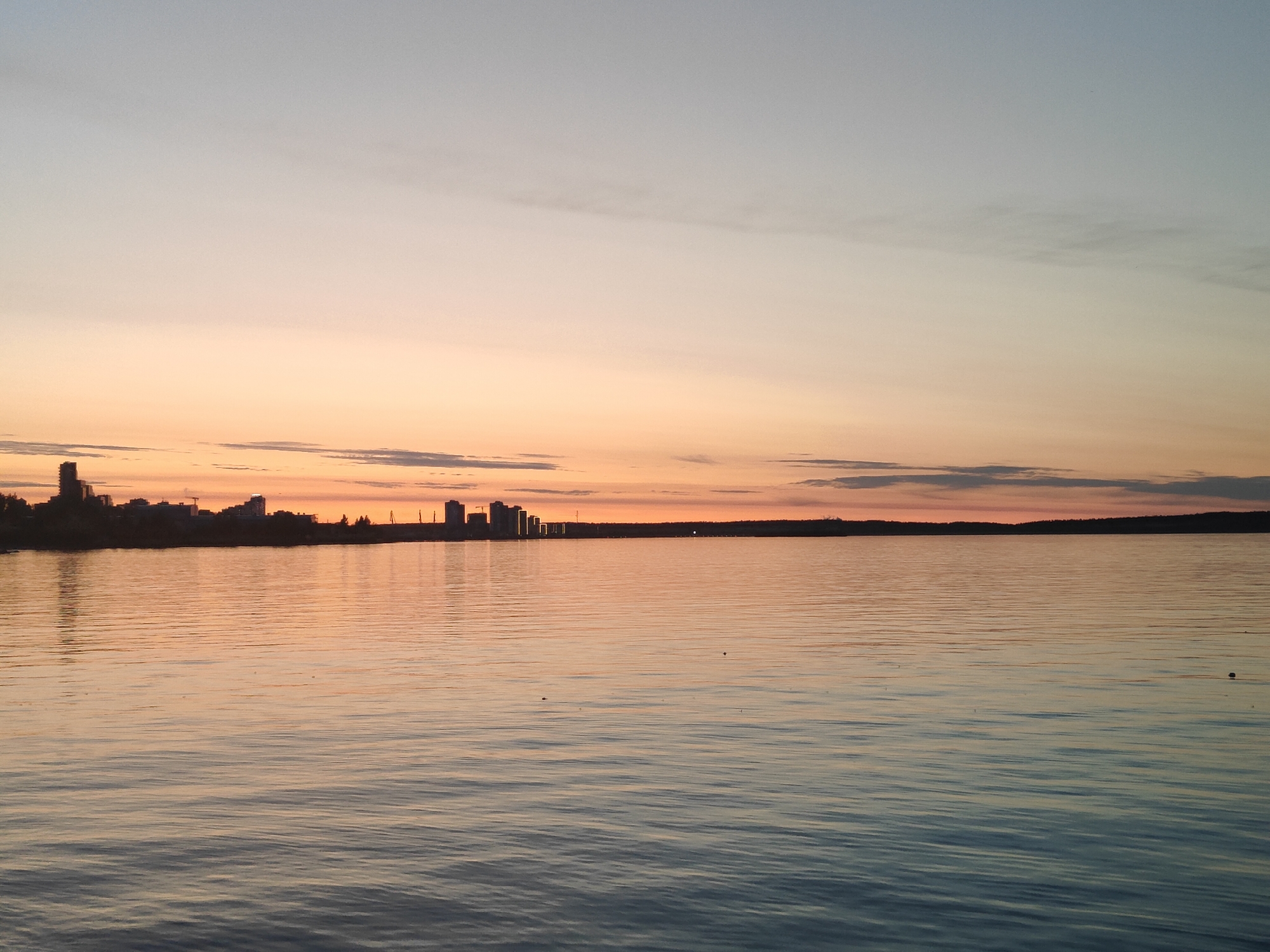 The image shows a serene sunset over a body of water, with a city skyline silhouetted in the distance. The sky transitions from a pale blue at the top to hues of orange and pink near the horizon, where wispy clouds are visible. The water is calm, reflecting the colors of the sunset, and ripples on its surface create subtle patterns. The city skyline features several tall buildings and structures, appearing as dark shapes against the vibrant sky. The overall scene is tranquil and atmospheric, suggesting a peaceful evening.