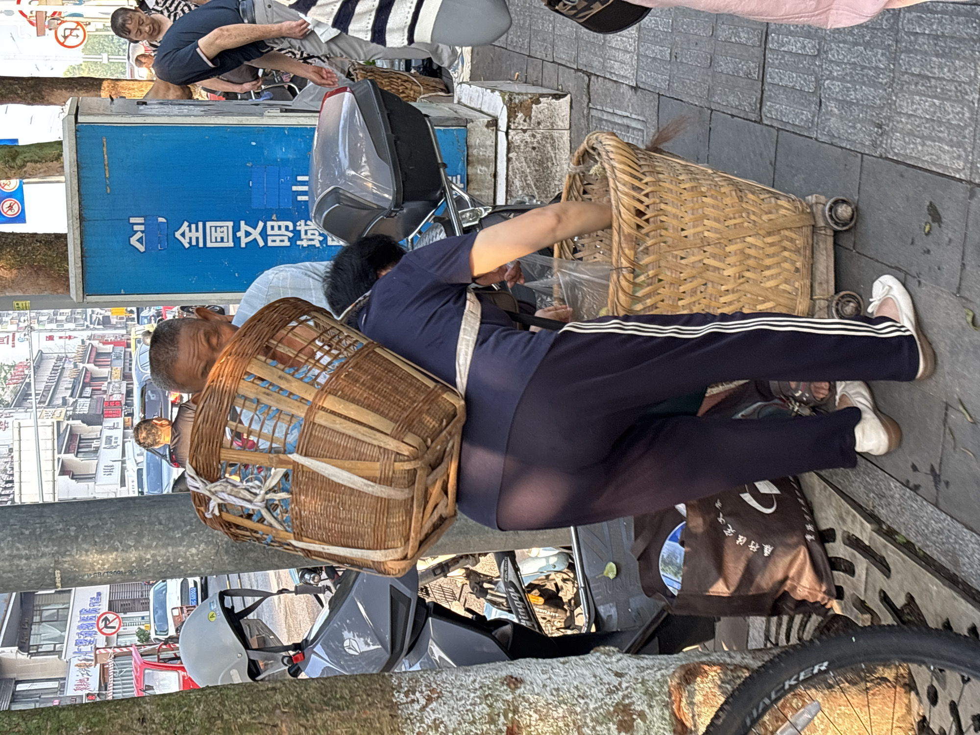 In the city of Jiangjin, China, on a sunlit sidewalk, a woman wearing a dark blue athletic suit with white stripes down the legs is positioned behind a large, wheeled wicker basket. She is bending down, reaching into a clear plastic bag within the basket. Strapped to her back is another, larger wicker basket, secured with white and blue straps. In front of her, a motorcycle with a grey helmet on its handlebars is parked next to a tree. Behind the woman, a blue utility box with Chinese characters is visible. To the right of the woman, another person, partially obscured, is wearing a striped shirt and grey pants. Further in the background, a red vehicle and other cars are present on a street lined with buildings.