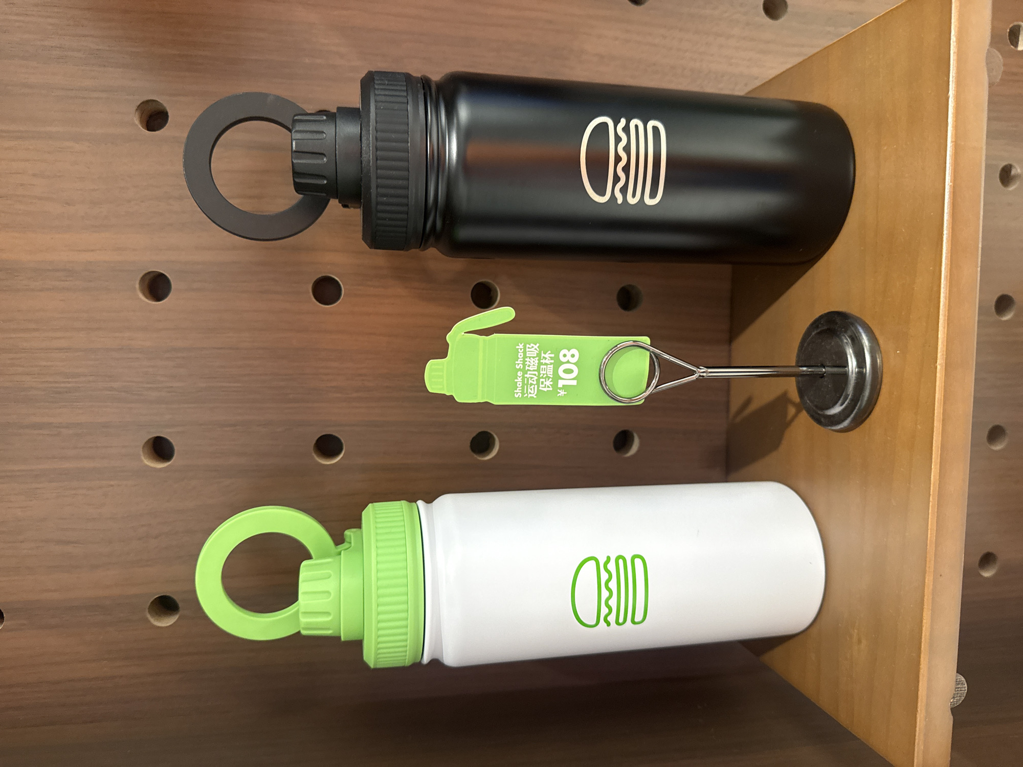 The image displays three water bottles on a wooden pegboard surface. The top bottle is black and has a white logo resembling a burger. It is attached to the pegboard by a black ring. Below it is a small, bright green water bottle or dispenser with text in Chinese characters and the number "108". This item is also attached to the pegboard by a metal loop. The bottom bottle is white with a green lid and a green burger logo similar to the top bottle. It is attached to the pegboard with a green loop. The overall setting appears to be a retail display or storage area, likely in Shanghai, China, given the location context. The lighting suggests an indoor environment. There are no people visible in the image, and no specific time of day or weather conditions can be inferred. The focus is on the display of the water bottles.