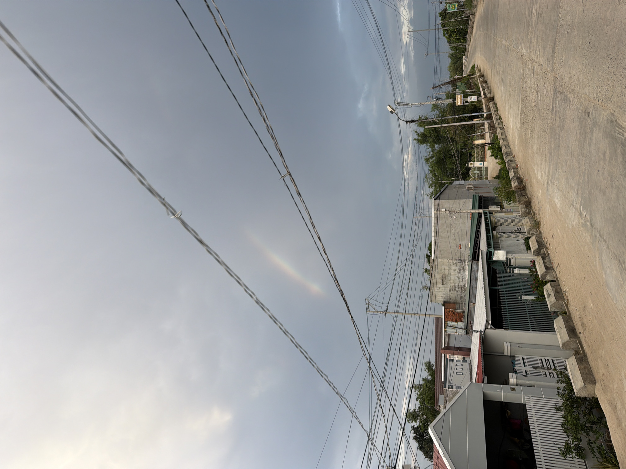 The image presents an ordinary street scene in Dong Hai Ward, Vietnam, captured on what appears to be an overcast day. The perspective looks down a concrete or asphalt road, lined with buildings and overhead utility lines.
The sky dominates the upper portion of the frame, showing a mix of light and darker grey clouds. A striking detail is a faint, small arc of color resembling a sun dog or a segment of a rainbow, visible in the central sky between the numerous crisscrossing power and communication wires. These overhead lines form a dense network across the frame.
Along the sides of the street are various buildings, likely residential homes or small businesses, characterized by concrete construction, metal gates, and utilitarian designs. Some buildings feature partial corrugated roofing. Green foliage from trees and bushes is visible behind and between the structures, adding natural elements to the urban setting. A modern streetlight is mounted on a pole on the right side of the road.
The street itself appears quiet and empty, with no people or vehicles visible, suggesting a calm moment. The time of day is daytime, possibly late afternoon, indicated by the soft, diffused light, yet bright enough to reveal the subtle atmospheric phenomenon.
Visible text on signs is mostly illegible; a green sign with faint white writing and a small white sign with red text (possibly numbers like "000") on a building are present but unreadable.