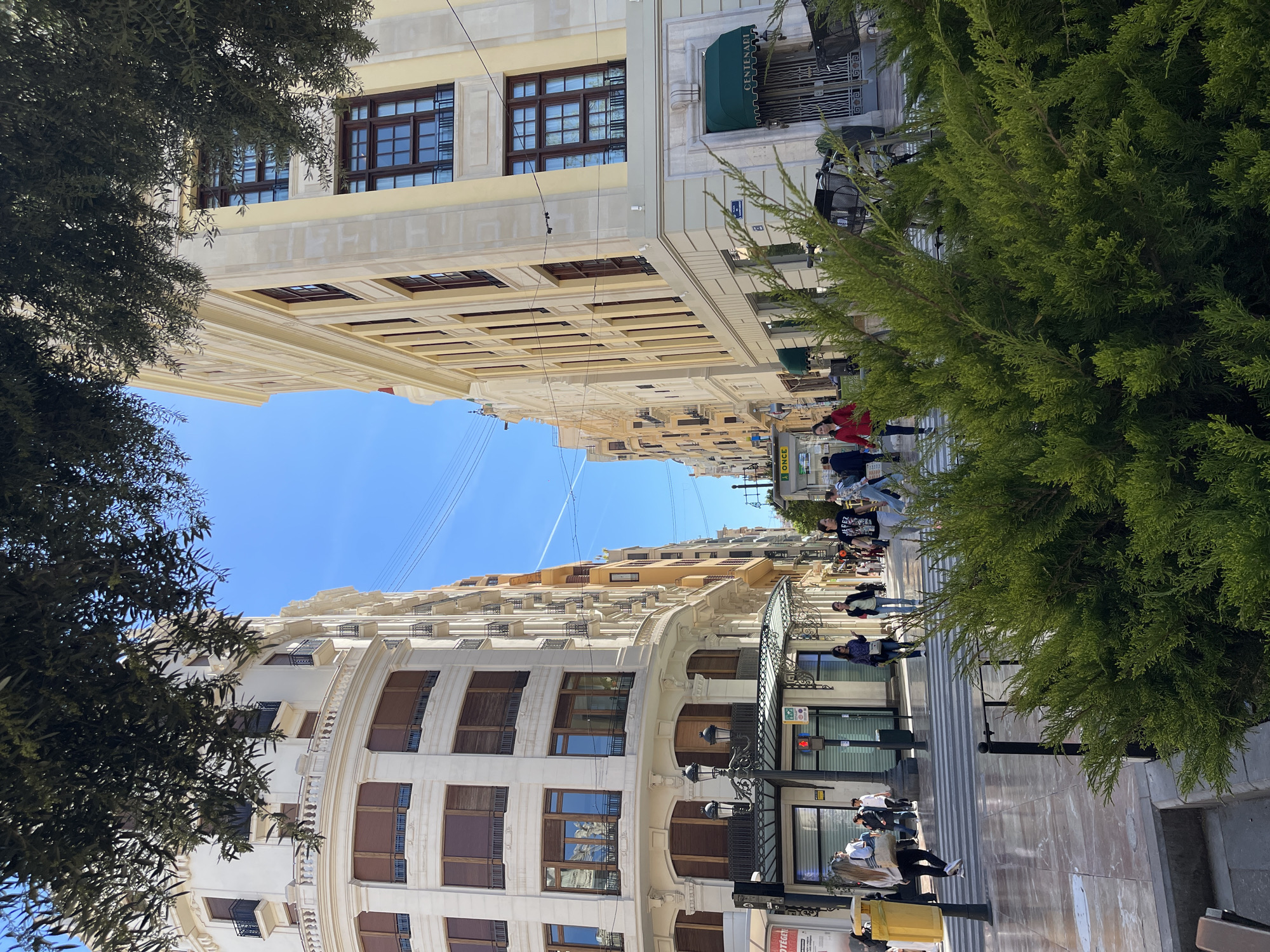 The image depicts a street in Valencia, Spain, specifically Carrer de les Barques near Plaça de l'Ajuntament. The perspective is from street level, looking upwards. Tall buildings line both sides of the street, with architectural details visible. The buildings are primarily white or light yellow with brown window frames.
Several pedestrians are walking along the street, including a group near a shop entrance. A large green tree stands on the right side of the frame, obscuring parts of the building facade. On the left is another tree.
Overhead power lines run across the sky between the buildings. Street furniture, including lampposts and signs, are also visible. The ground is paved and reflects light. The sky is clear blue.
