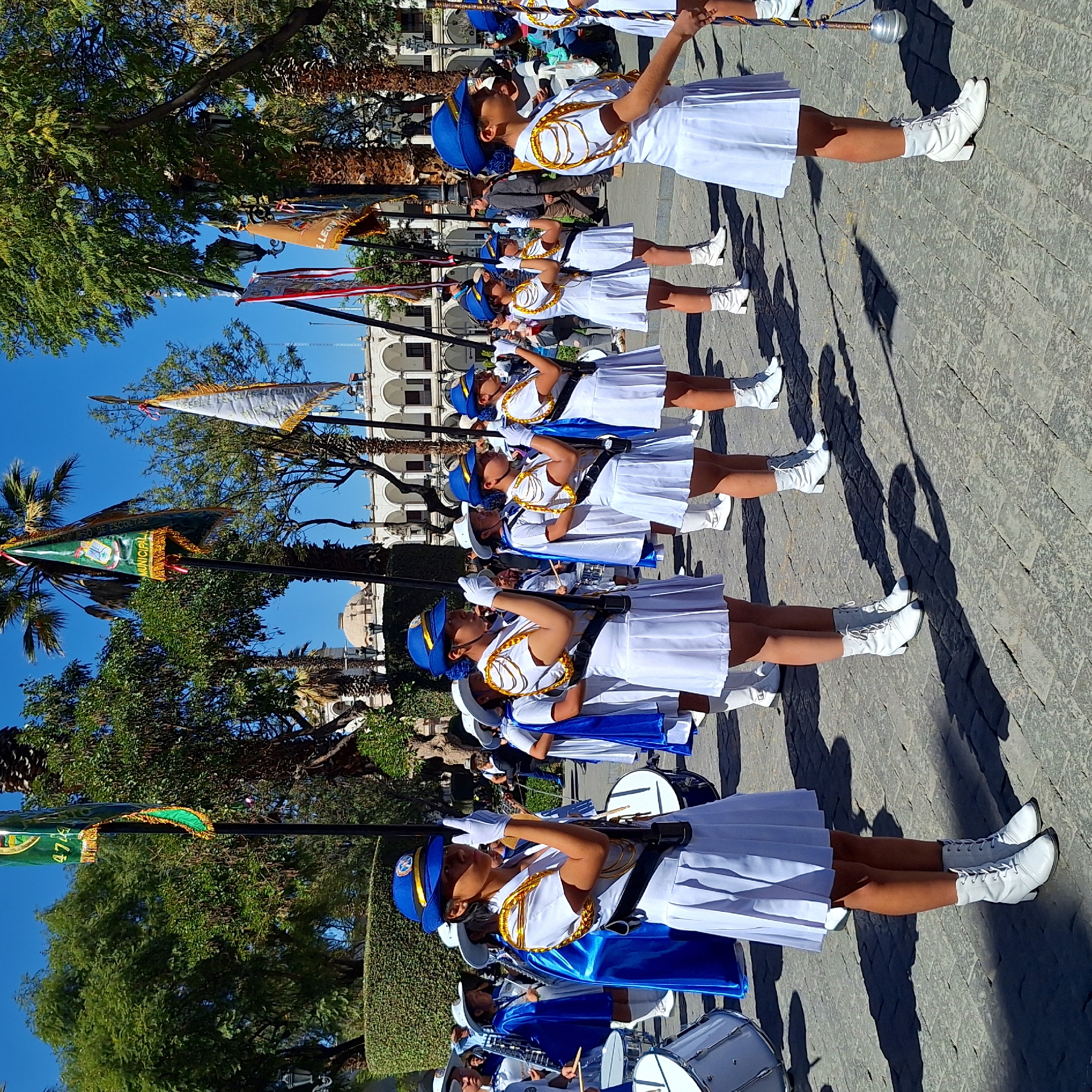 A line of young women in white uniforms with blue accents, including hats and capes, stand in formation on a paved street in Unknown, Unknown. They appear to be part of a marching band or color guard, as some hold flags or batons. Trees and buildings line the street, suggesting an urban setting. The sky is blue, indicating a sunny day.