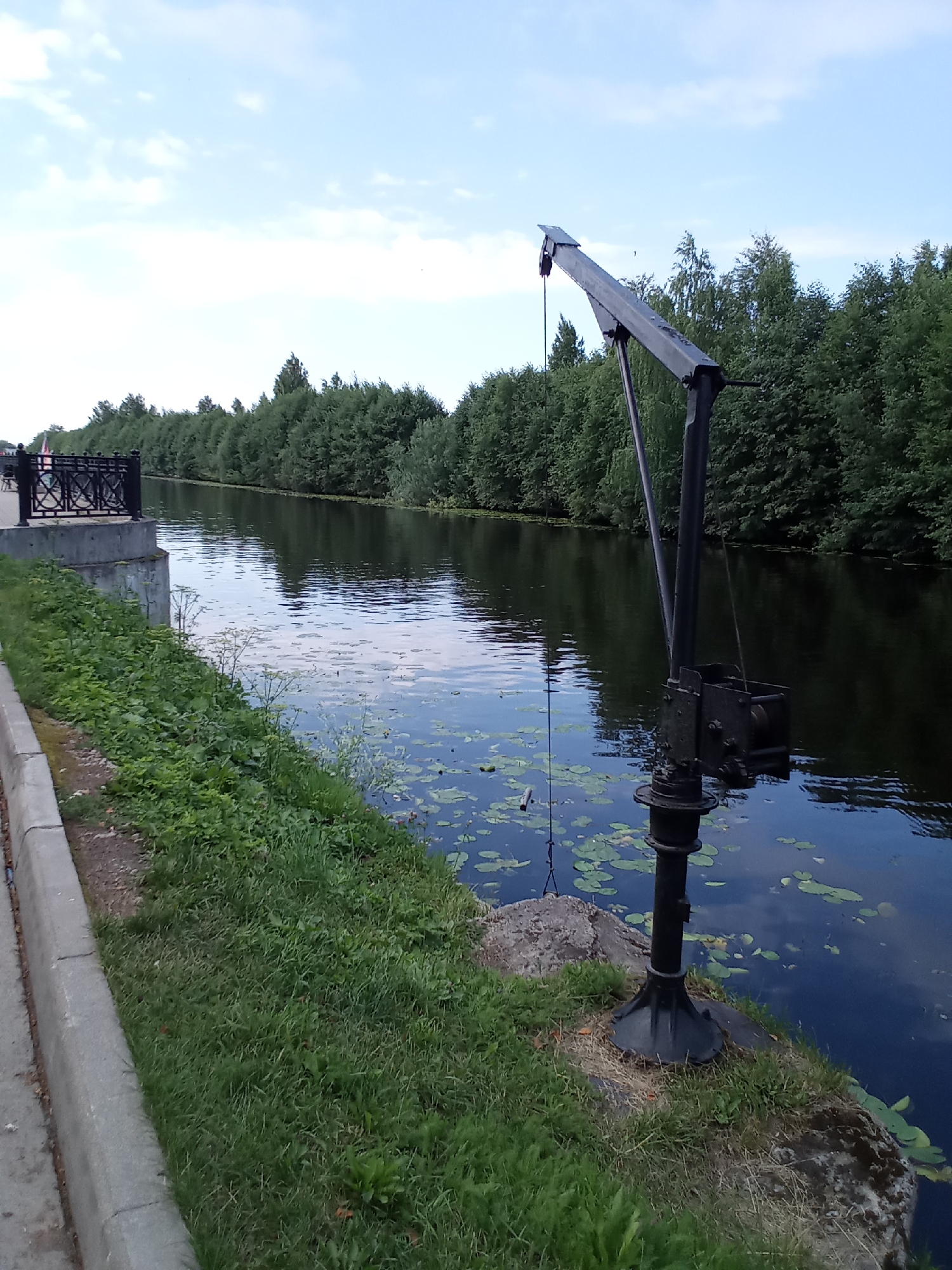 In Belozersk, Russia, a black metal crane stands on the grassy bank of a dark, tranquil canal. The crane's boom extends over the water, which is dotted with lily pads. A concrete walkway with a decorative metal railing runs along the left side of the canal, separated from the water by a strip of grass and vegetation. In the background, a dense line of green trees forms the opposite bank of the canal, under a pale blue sky with scattered clouds. The scene appears to be a peaceful, natural setting within the city, possibly a public park or embankment area.