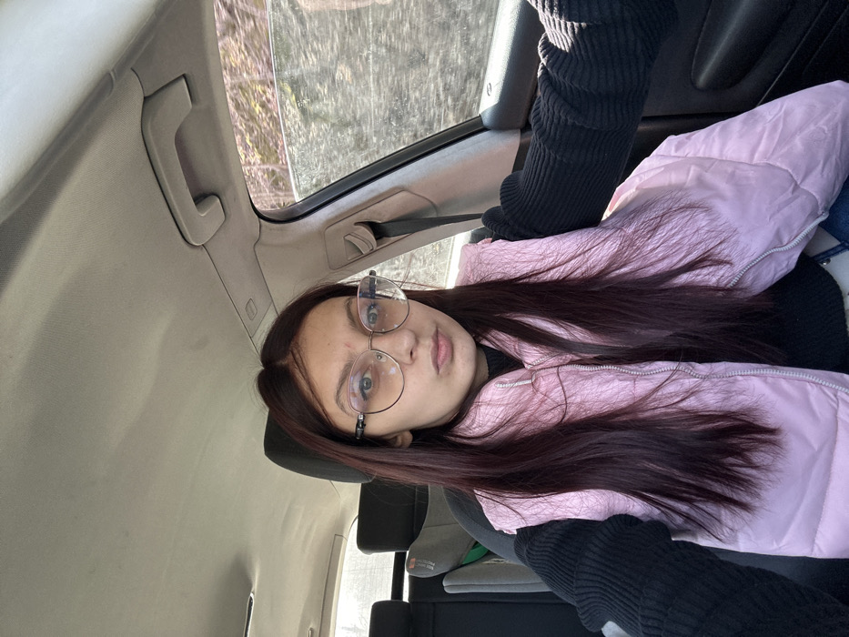 A young woman with long, dark reddish-brown hair, wearing rimless eyeglasses, a black ribbed long-sleeve top, and a light pink puffer vest, is seated in the front passenger seat of a car. She is looking directly at the camera with a neutral expression, her lips slightly pursed. A child car seat is visible in the rear passenger seat behind her. Through the car window on the right, blurred green and brown vegetation, likely trees or bushes, is visible, suggesting movement or a natural environment. The car's light-colored ceiling and a grey grab handle are visible above. The scene takes place in the city of Todoretsite, Bulgaria.