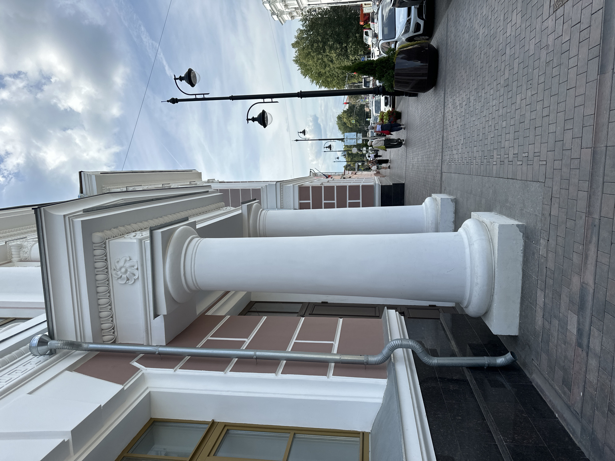 A photograph taken from a low angle, looking down a cobblestone sidewalk lined with white-columned buildings and a street with parked cars and pedestrians. The architecture suggests a historical European city, with ornate detailing on the buildings, such as cornices and decorative relief. The sidewalk is paved with rectangular cobblestones. Several lampposts are visible along the street. People are walking along the sidewalk in various directions. The sky is partly cloudy, with some scattered white clouds. The overall impression is a lively urban scene on a clear day. The specific location is not identifiable from the image alone.