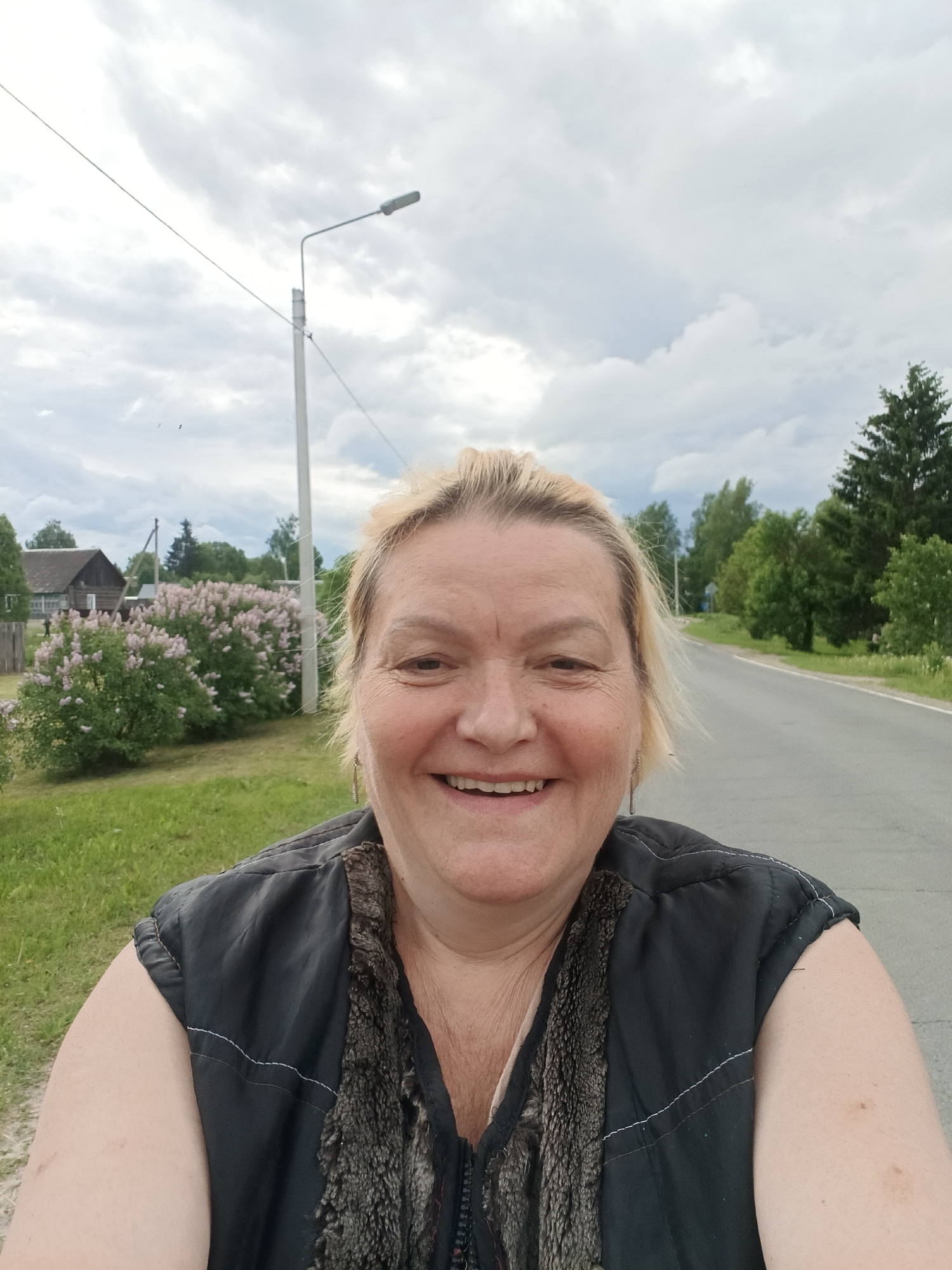 A woman is standing on a road in Красный Маяк, Russia. She is smiling at the camera. Behind her is a street lamp, a road, and trees. In the background is a wooden house with a lilac bush in front of it. The sky is cloudy.