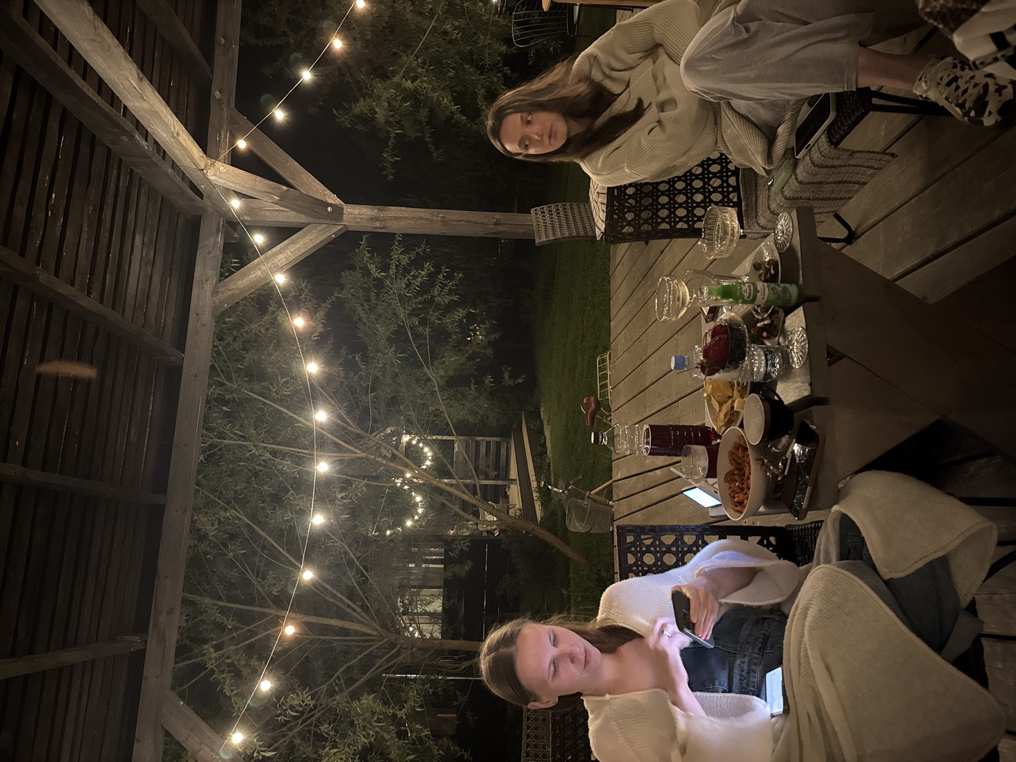 Two women are seated at a table laden with food and drinks, with festive string lights illuminating the surrounding outdoor space. The setting appears to be Prudki, Russia, possibly a garden or patio area. One woman is looking towards the camera while seated, while the other is looking down at a phone. The table contains various items including wine glasses, bottles, fruit, and snacks. A bicycle is also visible in the background. The scene is overall dimly lit but festive.