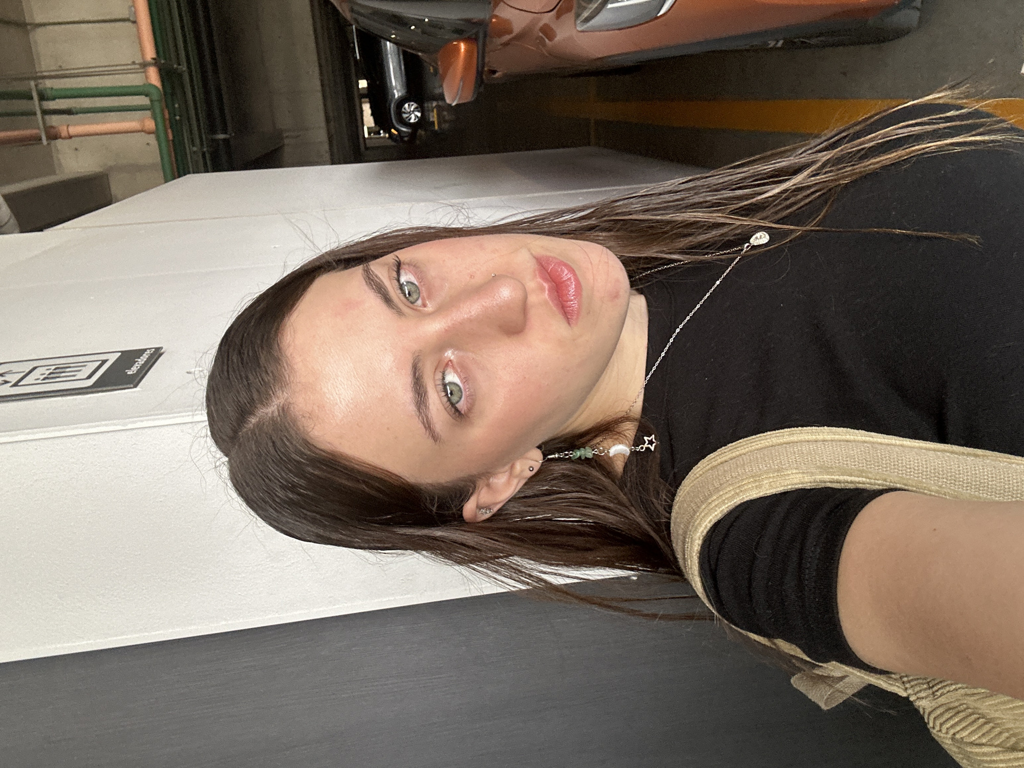The image is a tilted self-portrait featuring a young woman in what appears to be an indoor parking garage in Huixquilucan de Degollado, Mexico. The woman, whose head and upper torso are visible, has long dark brown hair with lighter streaks, parted on her left side (viewer's right). She has striking light green eyes, defined eyebrows, and is wearing pink lipstick. A small nose piercing is visible on her nostril.
She is dressed in a black top and wears a thin silver chain necklace with a small, star-shaped pendant. On her right ear (viewer's left), she has a unique dangling earring composed of green beads, a white crescent moon charm, and a silver wire, along with a small stud earring. A beige or gold woven strap, likely from a bag, is visible over her right shoulder. She looks directly at the camera with a neutral and composed expression.
The background elements confirm an indoor, industrial-style setting. A brown/copper-colored car is partially visible behind her head. To her left, green and orange pipes are visible running along a wall or ceiling. Below her, there's a white wall or structure, and a dark gray floor or wall. A yellow painted line, typical of parking spaces, is discernible on the ground behind her. A black rectangular sign with white text and a QR code-like symbol is visible on the white structure, displaying the word "electroleaves". The lighting is bright and even, consistent with a well-lit interior or daytime.