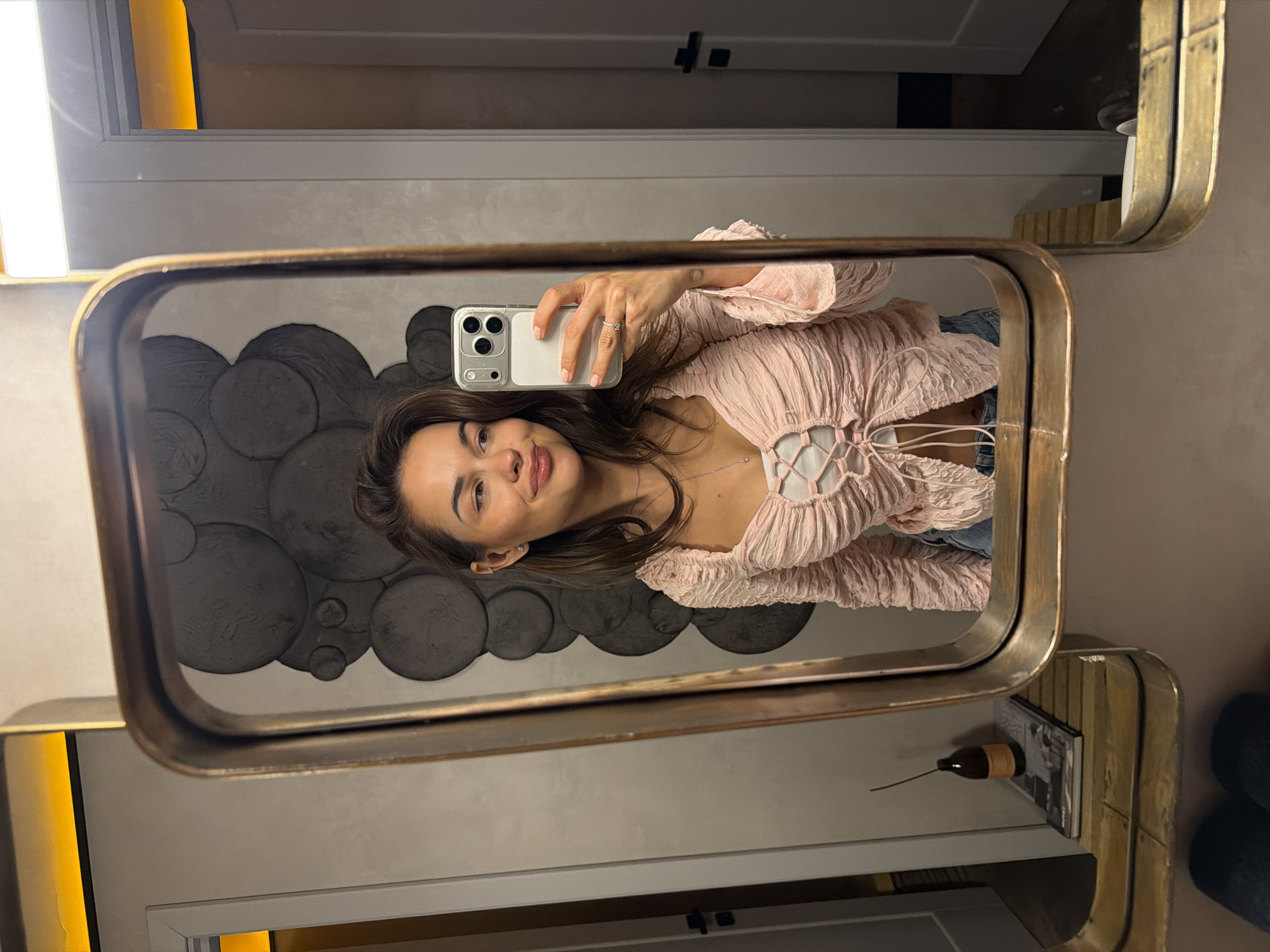 The image, rotated 90 degrees clockwise, presents a mirror selfie of a young woman in an indoor setting. She is reflected in a rectangular mirror with a worn, brushed golden or brass-colored frame. The woman has dark brown hair and eyes, a subtle smile, and is winking playfully with her right eye. She wears a light pink, textured top with a ruched, gathered design, featuring a criss-cross lace-up detail over a white inner layer. A delicate silver necklace adorns her neck, and a small stud earring is visible in her left ear.
She holds a silver-colored smartphone, likely an iPhone with multiple rear cameras, in a clear case with her right hand. A silver ring is visible on her right ring finger, and her fingernails are painted a light pink or nude shade.
The background reflected in the mirror showcases a dark gray wall with abstract, three-dimensional circular patterns of various sizes, creating a textured design. To the lower right, a shelf or counter holds a small, dark brown bottle with a narrow neck and a long stopper or applicator, placed alongside a stack of books or magazines. Above the mirror, a bright, rectangular light fixture and a soft golden-yellow light source are also reflected, indicating a well-lit indoor environment. No legible text is visible within the image.