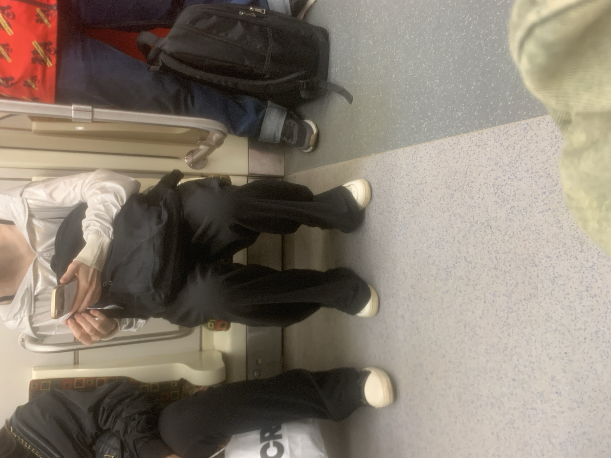 The image depicts the interior of a public transport vehicle, likely a metro or train car, in Saint Petersburg, Russia. Several passengers are visible, mostly from the waist down, engaged in typical commuting activities.
At the top of the frame, a person is seated, wearing dark blue jeans and a red garment with repeating yellow patterns. A black backpack rests on the speckled grey floor next to their dark-colored sneakers. In the middle of the frame, another passenger is seated, dressed in a white long-sleeved top and black wide-leg trousers. They are holding a light-colored smartphone with both hands, appearing to interact with it. Their feet are clad in white sneakers. Further down, at the bottom of the frame, a third person is partially visible, also seated, wearing black trousers and white sneakers. A white bag with the black text "CR" is placed near their feet. On the far right, a blurred section suggests another person's leg or foot.
The setting is characterized by light grey speckled flooring and upholstered seats featuring a geometric pattern in red, yellow, and black hues. Silver metal handrails are installed along the seating area. The lighting is bright and even, consistent with artificial illumination inside a public conveyance. No specific time of day or weather conditions are discernible. The overall scene captures routine urban travel.