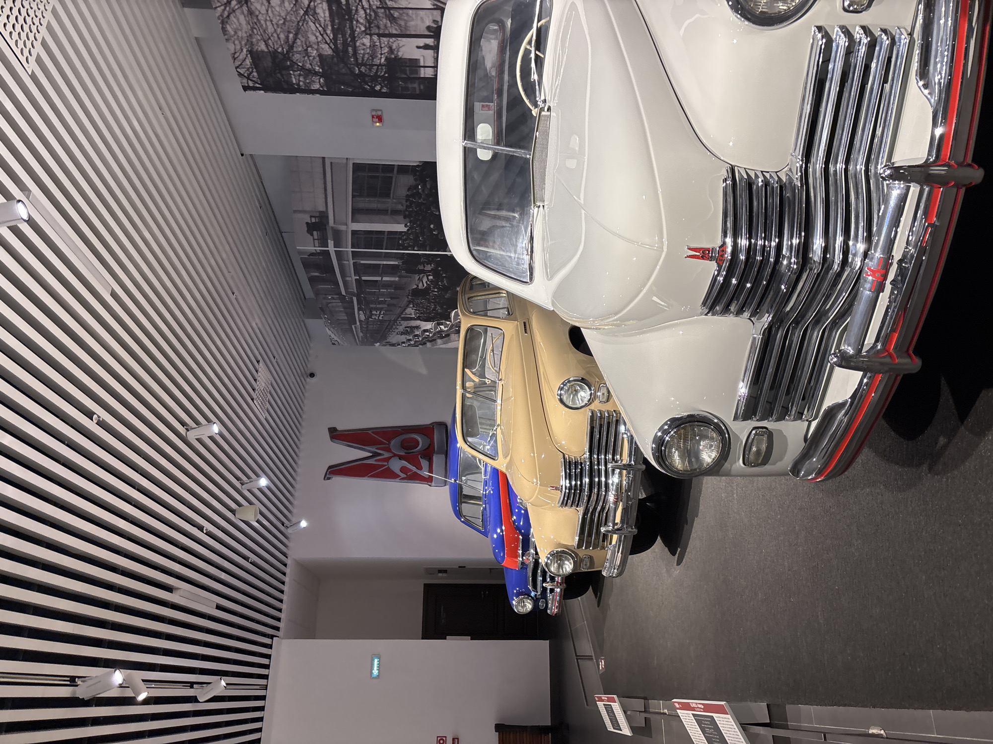 The image captures a display of vintage automobiles inside the Museum of Automotive Technology in Verkhnyaya Pyshma, Russia. Three cars are prominently featured in a line, viewed from the front. The closest car is a white sedan, followed by a tan sedan, and then a blue sedan with a red stripe. All vehicles appear to be from the mid-20th century. The museum environment includes a modern ceiling with slatted panels and integrated lighting, and a large black and white mural on the wall behind the cars depicting a historical scene of a factory or industrial building with a crowd of people. A stylized red logo with the number "20" is visible on the wall above the blue car. The floor is dark and appears to be a matte material. Information displays are visible to the left of the cars. There are no people visible in the immediate foreground or interacting with the cars. The overall impression is a well-maintained museum exhibit showcasing historical vehicles.