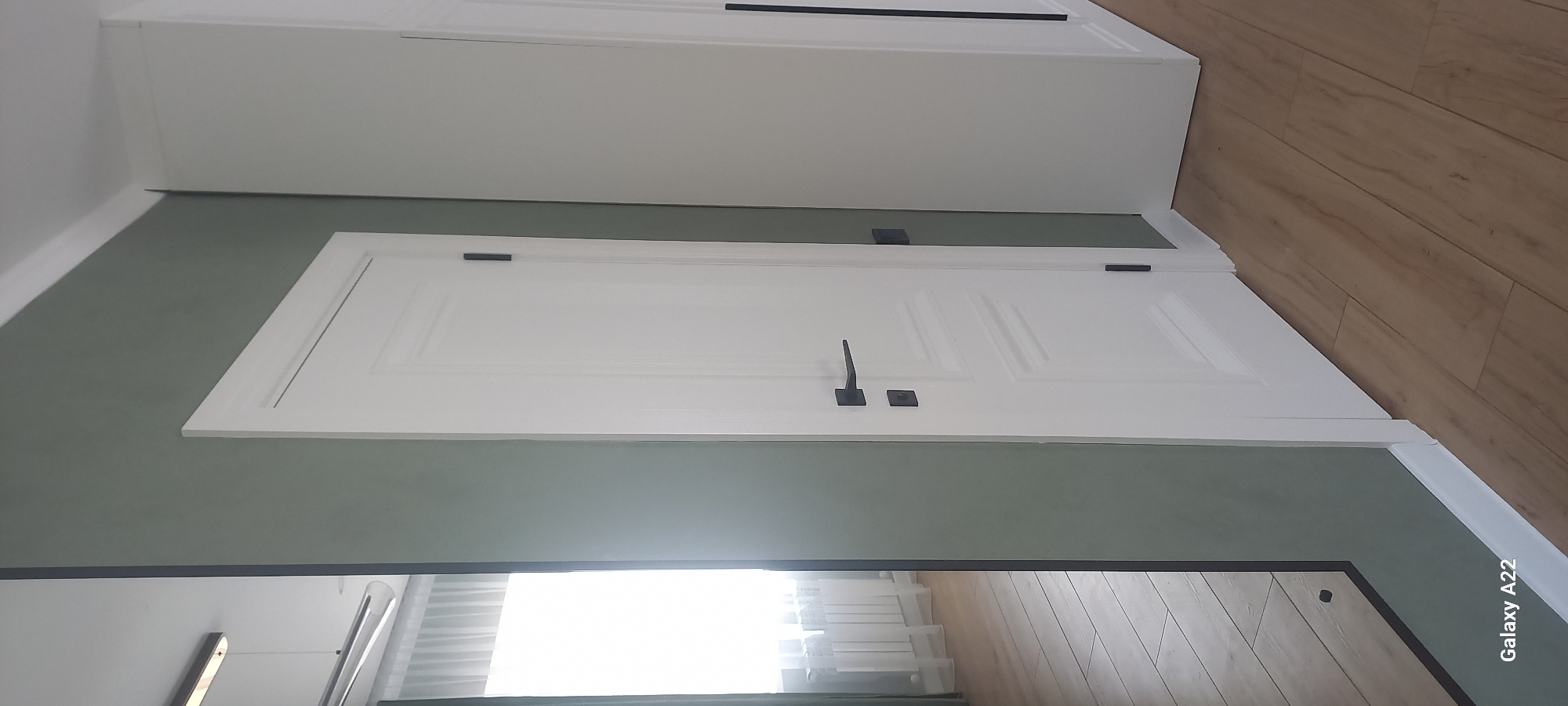The image depicts an interior scene, likely a hallway or a room with doors. The primary subjects are white doors with dark hardware, including handles and hinges. One door, in the foreground, has two panels and a distinctive L-shaped handle. Another door, further back and higher up, also appears to be white with dark hardware. The walls are painted in two distinct colors: a muted green on the lower section and white on the upper section, which also features part of a white cabinet or built-in furniture. The floor is made of light brown wooden planks laid diagonally. The lighting suggests it is daytime, with natural light illuminating the space from an unseen source, possibly a window or doorway to the right. The overall impression is one of a clean, modern, and well-maintained interior. The text "Galaxy A22" is visible in the lower right corner of the image, indicating the device used for capture. There are no people or animals visible in the scene. The location context is given as Oral, Kazakhstan.