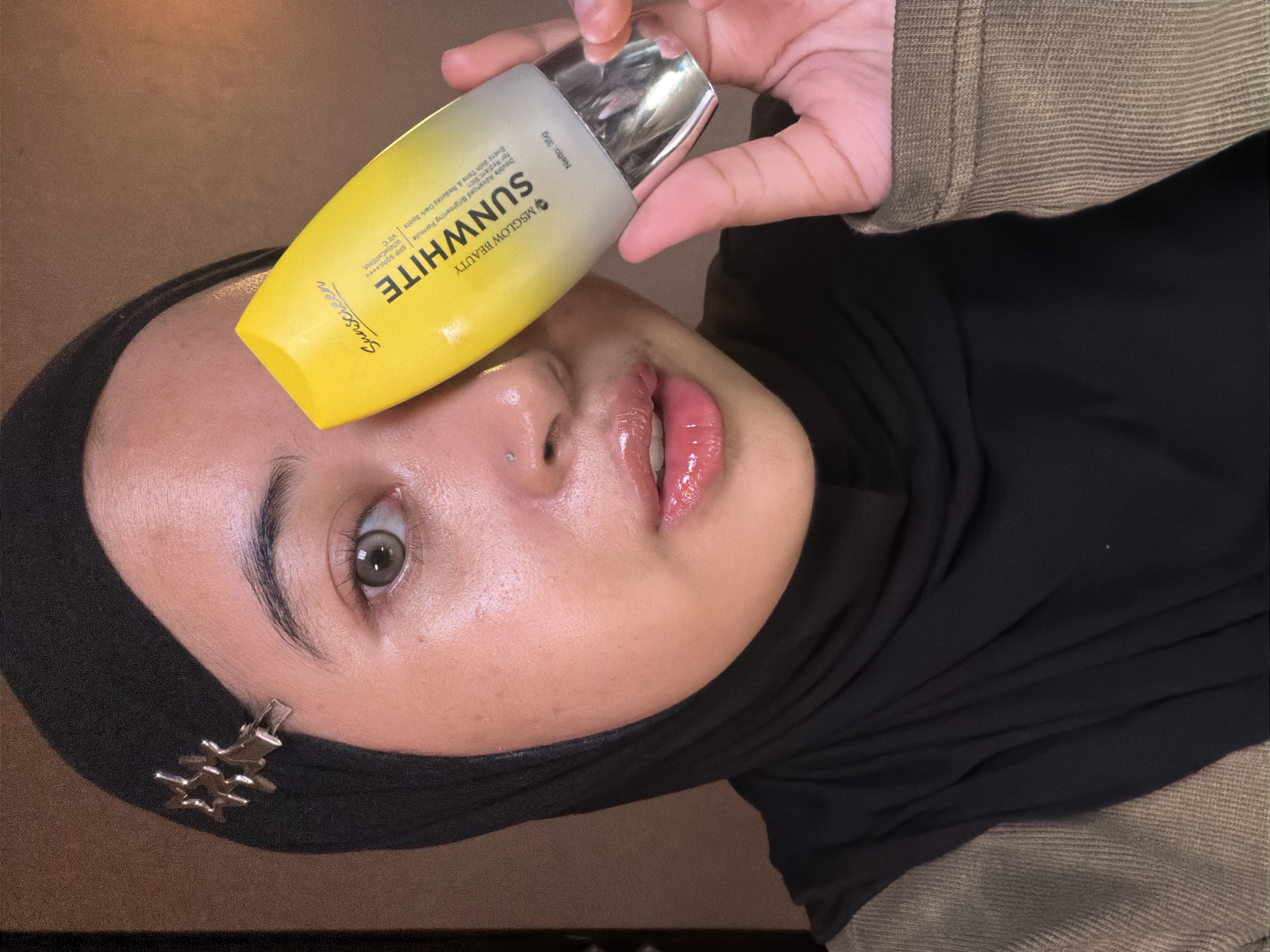 The image captures a close-up, angled shot of a person, likely a woman, with medium skin tone, holding a product bottle above her face. She is wearing a black hijab, secured with a small silver star-shaped clip near her temple, and a greenish-brown ribbed long-sleeved top. Her face is well-lit, with a slight sheen on her forehead and nose, and her lips are glossy pink. She has a small silver stud piercing on her nose and one eye is visible, looking towards the camera.
The central object is a yellow and silver sunscreen bottle, held by her right hand just above her forehead. The bottle's label is clearly legible, displaying brand and product information. Visible text on the bottle includes "Sunscreen", "MSGLOW BEAUTY", "SUNWHITE", and details like "Double Advanced Brightening Formula for Radiant Skin", "Evens Skin Tone & Reduces Dark Spots", "SPF 50 PA++++", "WhiteCellDNA", and "Vit C". The net weight "Netto: 35g" is also visible.
The background is dark and largely indistinct, suggesting an indoor setting with low ambient light, possibly illuminated by a direct light source highlighting the subject and product. The overall impression is a posed shot for product promotion. The location context provided is East Jakarta, Indonesia.