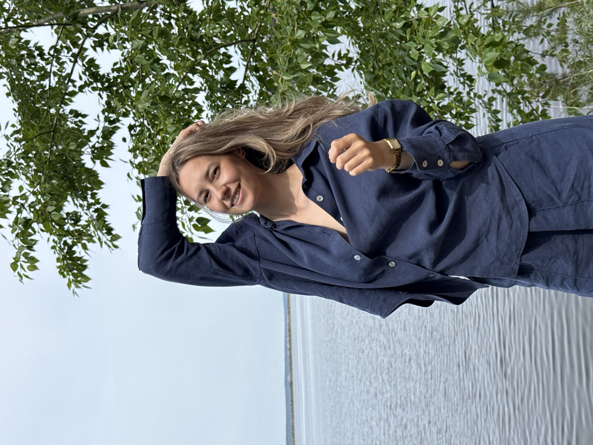 A woman with light brown hair stands in an outdoor setting with a body of water, possibly a lake, in the background. She is wearing a navy blue linen suit, consisting of a button-down shirt and pants. She has a gold watch on her left wrist and a smile on her face. Her left hand is raised, touching her hair. The setting includes green trees and foliage above and to the left of the subject. The location is likely Chaikovsky, Russia.