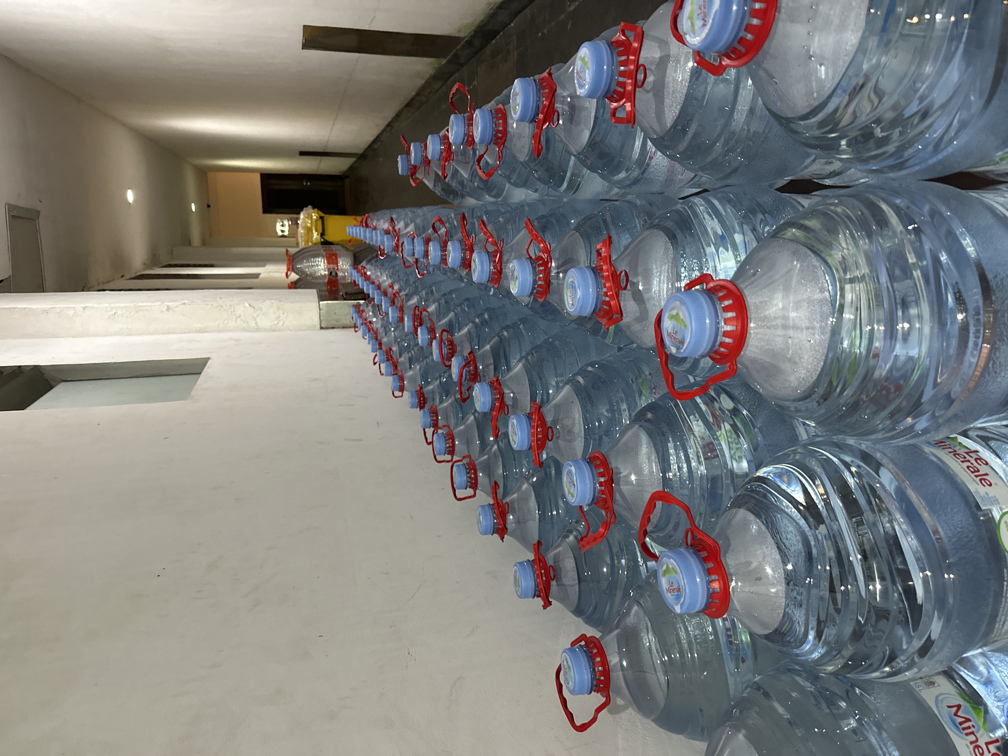 A long, narrow hallway, likely in a commercial building or warehouse, is lined with rows of large, clear plastic water bottles. The bottles are filled with water and capped with blue lids and red handles, suggesting they are for sale or distribution. The hallway is illuminated by overhead lighting, casting shadows and highlighting the uniformity of the bottled water. The walls are painted a light color and appear somewhat aged. In the background, the hallway recedes into the distance, with a dark doorway visible at the far end. There are no people visible in the image. The scene is clean and orderly, indicating a storage or staging area. The location is Kuta Selatan, Indonesia.