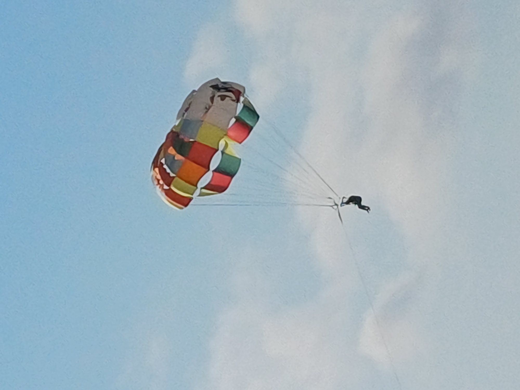 A person is parasailing against a backdrop of a pale blue sky with scattered clouds. The parasail is a large, colorful canopy with a patchwork of squares in red, orange, yellow, green, and white. It is inflated and catching the wind, pulling the person along. The person, dressed in dark clothing, is suspended beneath the parasail by tethers. Their body is angled, suggesting movement through the air. The scene evokes a sense of outdoor recreation and exhilaration. The bright colors of the parasail contrast with the soft hues of the sky, indicating it is likely daytime, possibly late morning or afternoon given the bright, even lighting. The image captures a moment of adventure and freedom. There is no visible text in the image.