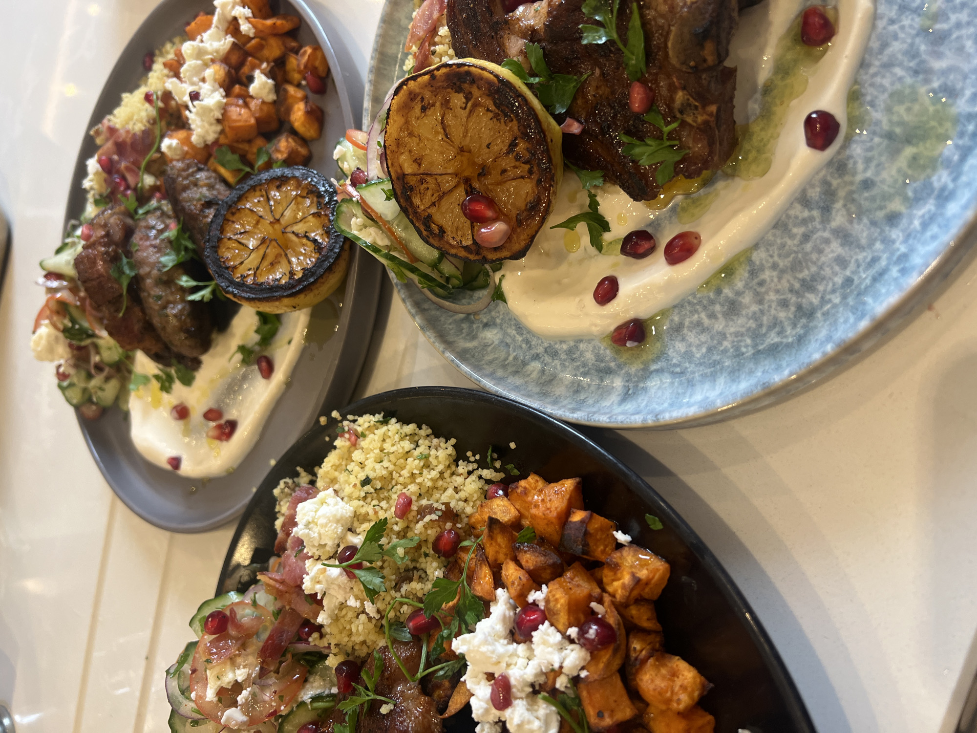 The image shows two plates of food, viewed from above. The first plate, on the upper right, contains lamb chops with a grilled lemon half, a dollop of white sauce, and pomegranate seeds. The plate has a blue pattern. The second plate, on the lower left, contains roasted sweet potatoes, couscous, feta cheese, and a salad of cucumbers, tomatoes, and red onions. The plate is black. The setting appears to be a restaurant or kitchen, with a white countertop. The precise location in London, United Kingdom cannot be determined from the image.