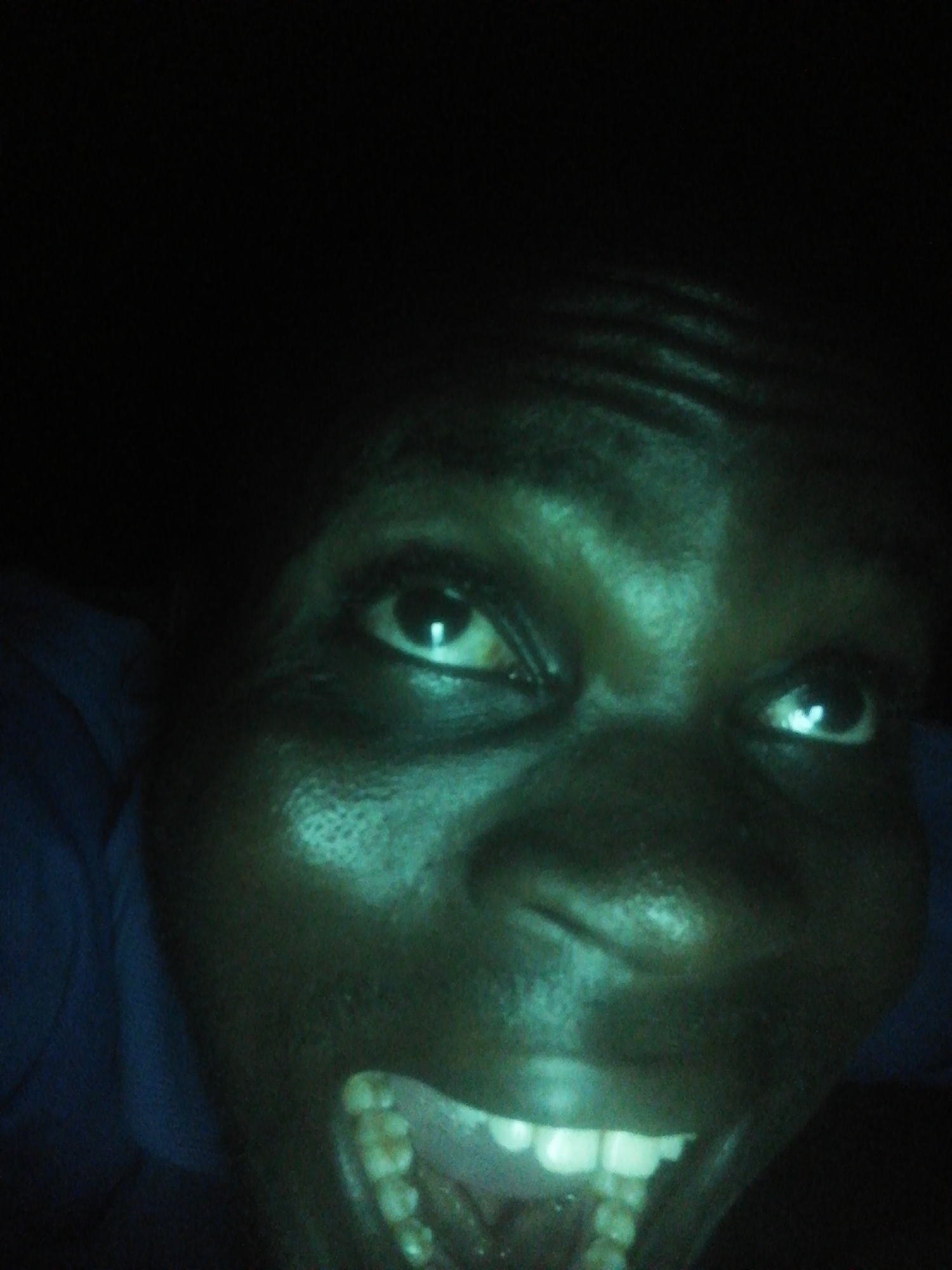 A close-up, low-angle photograph shows a person's face illuminated with a greenish hue against a dark background. The person's mouth is open, revealing their teeth. The person's eyes are open and directed upward. The location and city are unknown.