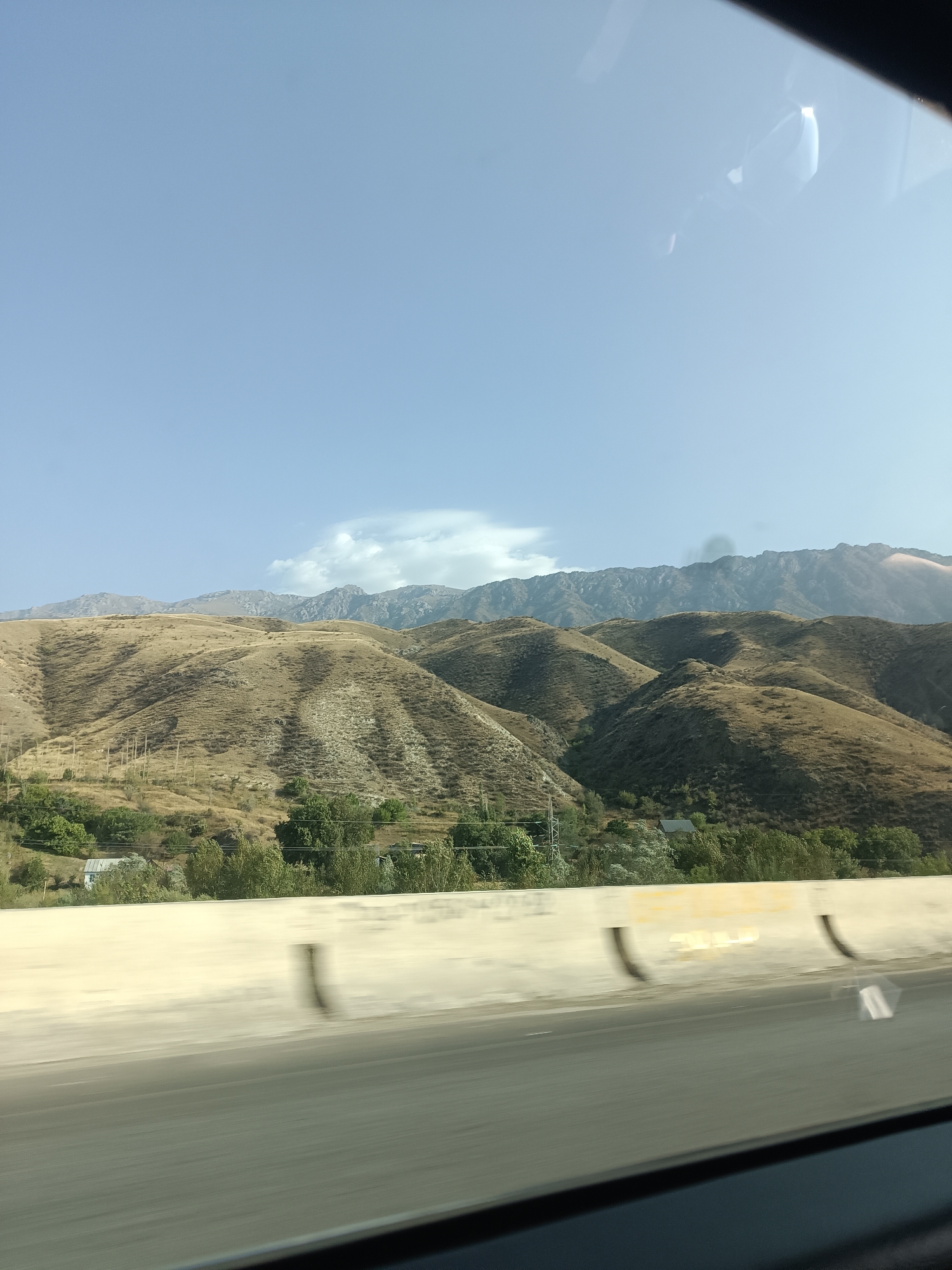 The image depicts a scenic view of rolling, dry, grassy hills under a clear blue sky with a few scattered clouds. The hills are covered in sparse vegetation, giving them a golden-brown hue. In the middle ground, a cluster of green trees and a small building are visible, suggesting a rural or semi-rural settlement. A concrete barrier, likely a roadside divider, is in the foreground, with a road surface blurred by motion. The background shows a range of rugged, distant mountains. The overall impression is of a vast, natural landscape. The location is identified as the city of Kara-Köl, Kyrgyzstan.