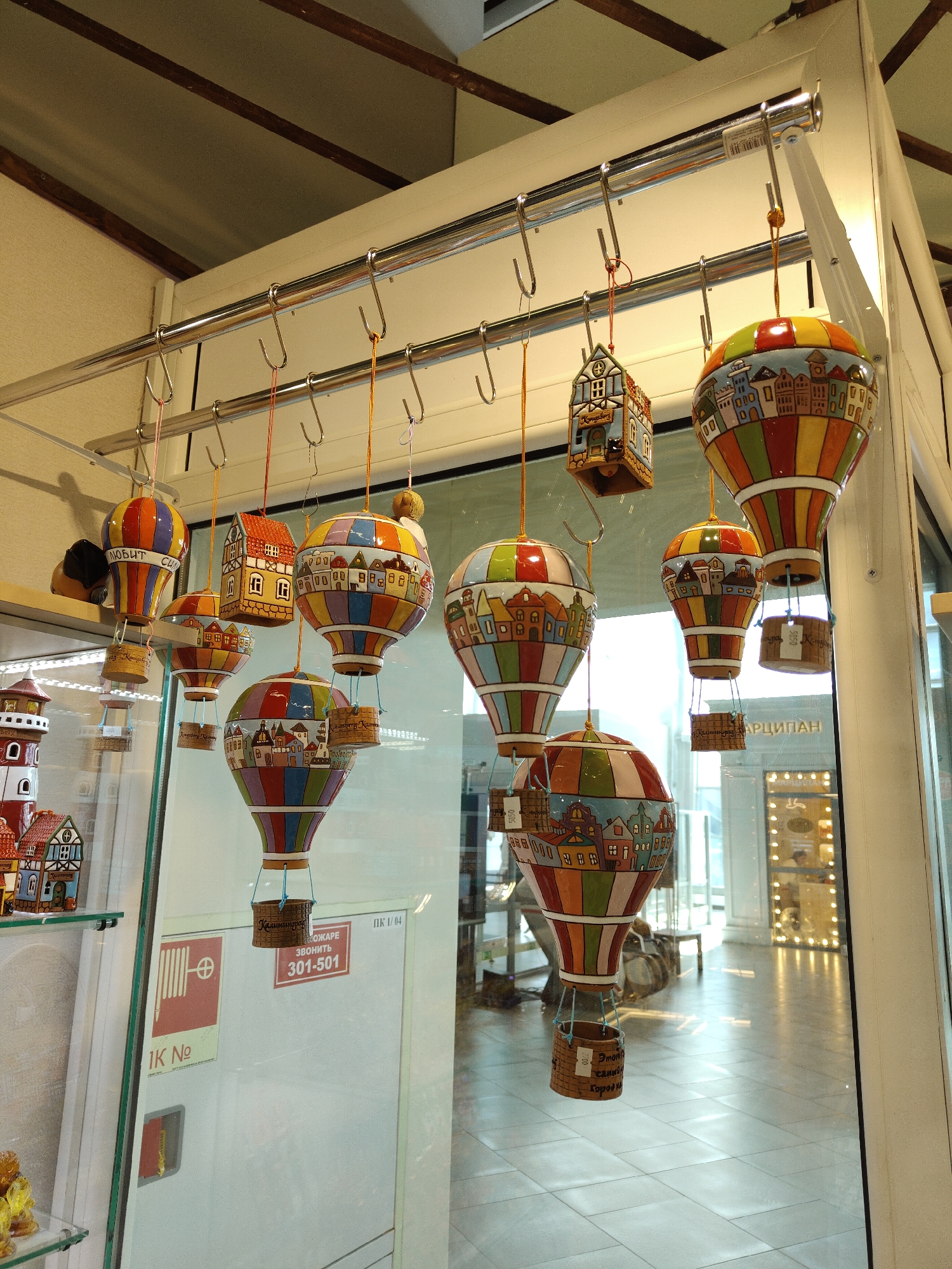 This is a close-up, eye-level shot of several decorative ceramic hot air balloons and houses hanging from hooks on a metal rod. The objects are brightly colored with intricate patterns depicting small towns. Some of the balloons have small baskets suspended from them, also adorned with decorative elements and text. The backdrop suggests an indoor setting, possibly a shop or market, with a glass partition reflecting a tiled floor and further displays in the background. The overall impression is one of a whimsical, handcrafted display. The signage visible on the glass partition, "301-501," and the Cyrillic text on some of the hot air balloon baskets, such as "Калининград" (Kaliningrad), indicate the location is likely Khrabrovo, Russia, a settlement near Kaliningrad. No people are visible in the immediate scene.