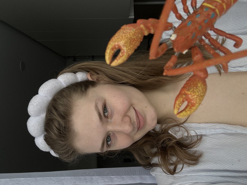 A fair-skinned woman with light brown, wavy hair smiles at the camera. She wears a fluffy, white headband. Positioned near her head is an orange toy lobster. She is wearing a striped shirt. The background consists of a wall on the left and a partially visible window or curtain below.
