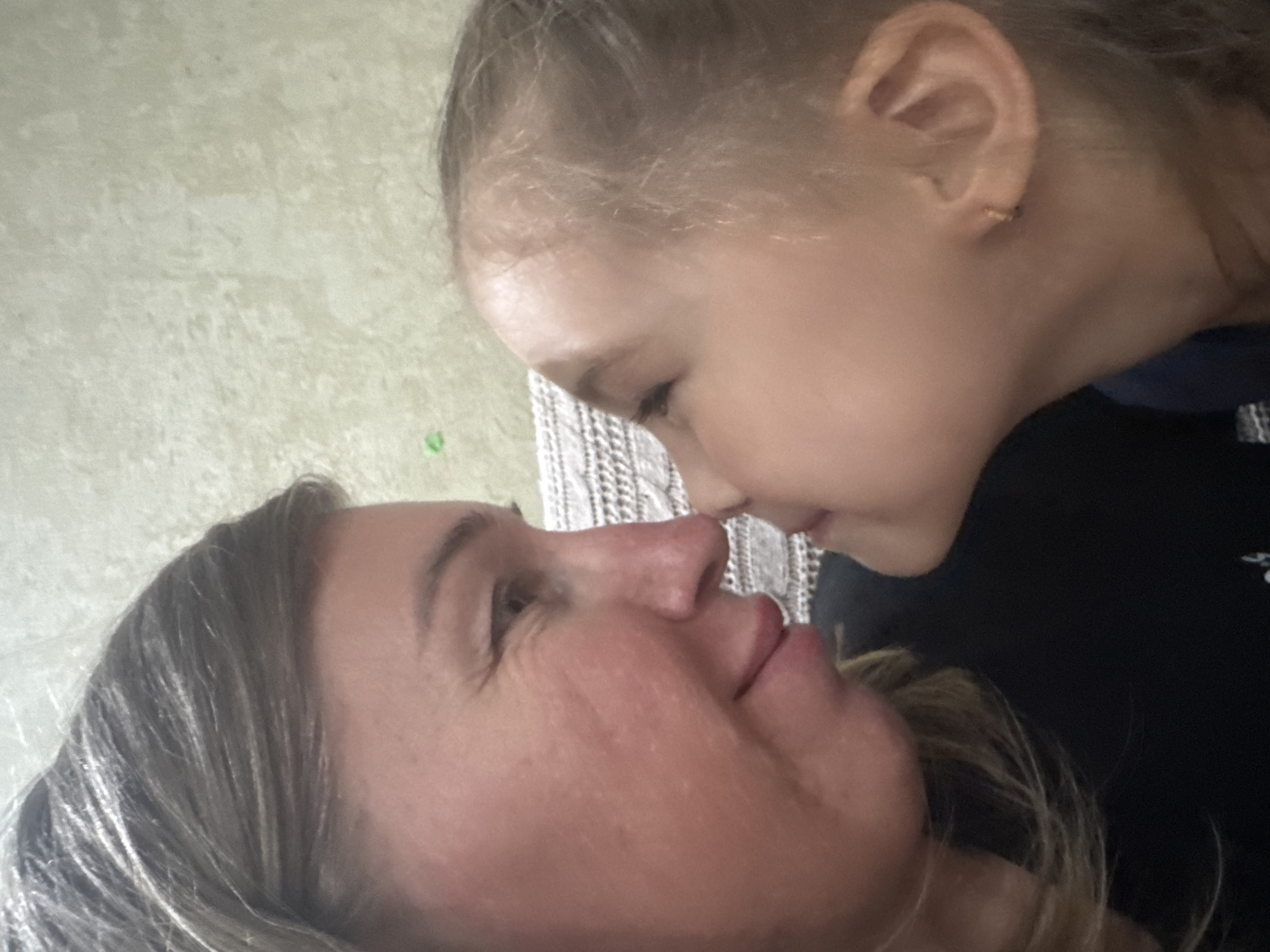 A close-up, candid photograph features a woman and a child in an intimate moment. The woman is positioned in the lower left of the frame, facing upwards with her eyes looking towards the child. The child, with lighter blonde hair, is positioned above and to the right of the woman, their faces close together. The subjects appear to be touching noses. The woman's skin appears red. The background is indistinct but seems to be an interior space with textured wallpaper. There is no definitive evidence within the image to confirm or deny the location of Tashkent, Uzbekistan.