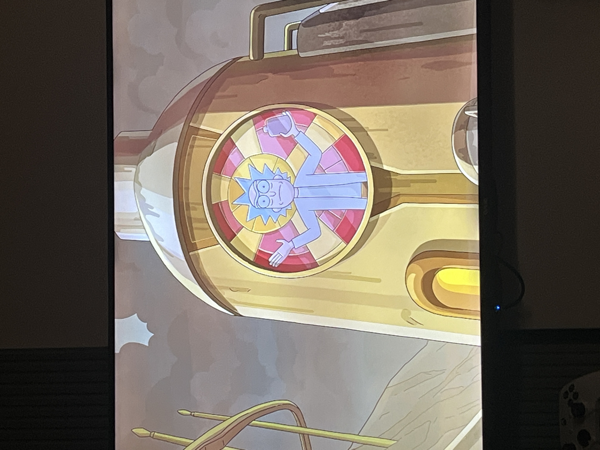 This media file shows a television screen displaying a cartoon image. The main subject is a character resembling Rick Sanchez from the animated series "Rick and Morty." He is depicted in a circular window with a stained-glass-like design featuring segments of yellow, pink, and red. Rick, drawn in a simplified, stylized manner with blue skin and spiky hair, is holding a purple rectangular object, possibly a flask or phone, in his right hand and has his left hand extended outwards.
The circular window is set within a large, metallic, cylindrical object that is predominantly golden or brass in color. This object appears to be part of a larger machine or vehicle. The background of the image shows a cloudy, grayish sky and some indistinct brownish structures or terrain below. There are also some yellow curved structures in the foreground. The overall aesthetic is that of a science fiction cartoon, with a vibrant and somewhat surreal design.
There is no visible text in the image. The image quality suggests it might be a photograph of a screen, with some glare and slight distortion. The lighting appears to be from the screen itself, illuminating the cartoon image. The overall mood of the depicted scene, based on the character's expression and posture, is one of casual confidence or perhaps mild bemusement. Given the context of the "Rick and Morty" animation style, the scene likely takes place in an extraterrestrial or interdimensional setting. The specific location in Krasnoyarsk, Russia, is external context and not visually represented in the image.