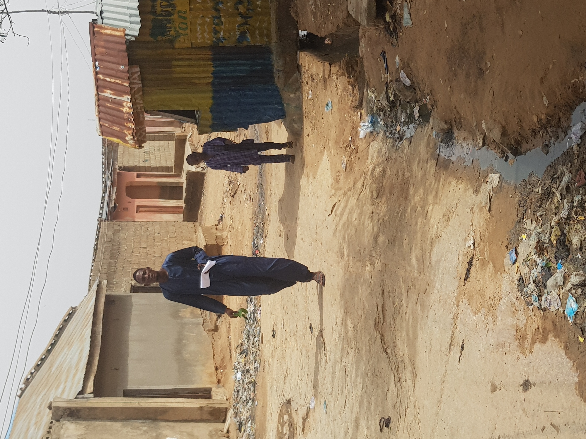 The image depicts a street scene in Dabawa, Nigeria. Two men are present. The first man wears a blue robe and holds a piece of paper and a green object in his hands. The second man, located further down the street, is wearing a plaid shirt. The street itself is unpaved and appears to be made of dirt. A drainage ditch filled with trash and waste runs along one side of the street. Buildings with corrugated metal roofs and unfinished exteriors line the street. Overhead wires are visible against the sky.