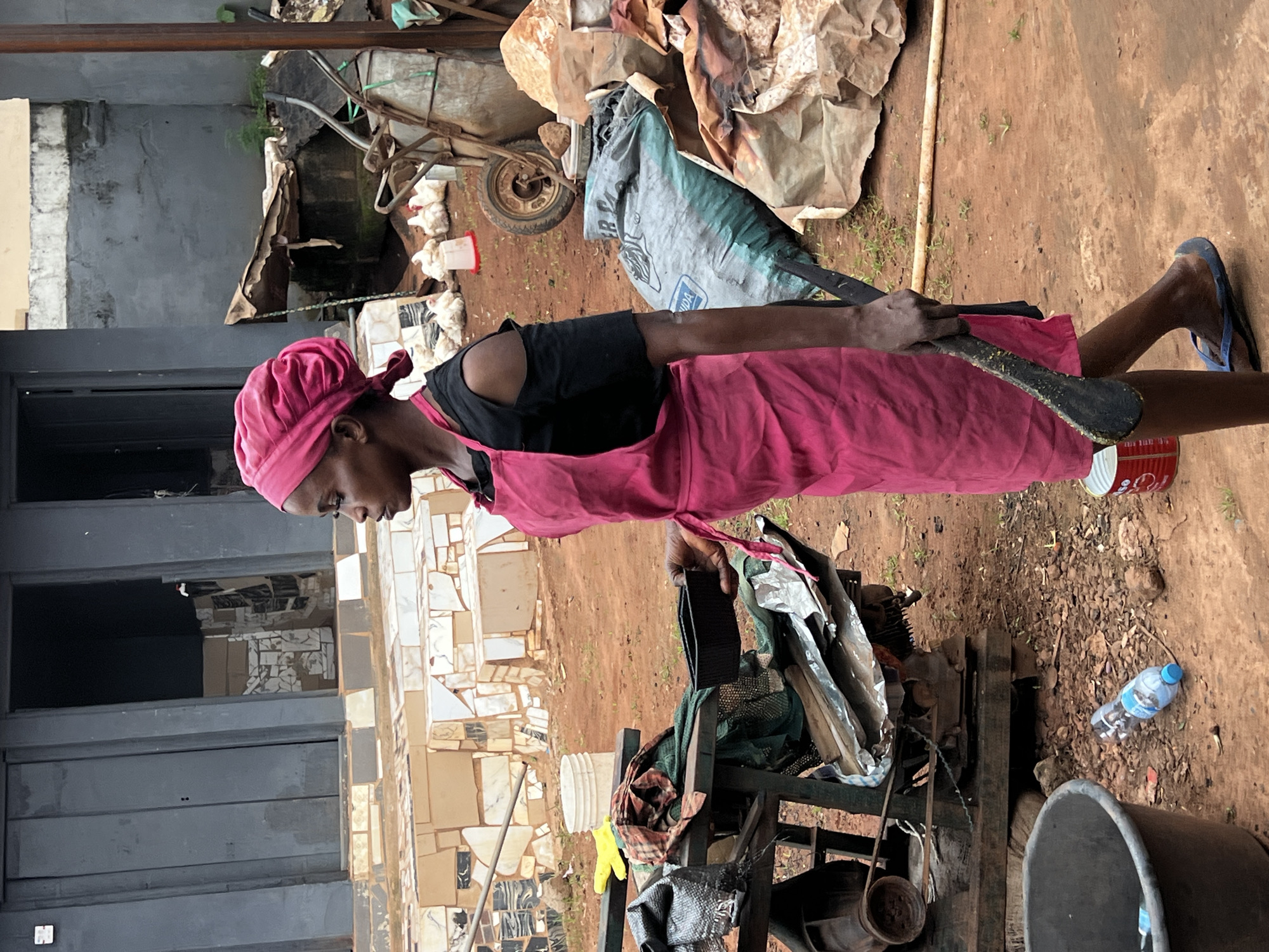 A woman wearing a pink headwrap and a pink apron over a black off-the-shoulder shirt walks outdoors in Nkpor, Nigeria, holding a large black utensil. She is looking to her left, with her body angled towards the right of the frame. Behind her, to her left, is a building with dark gray doors and a tiled facade. In the middle ground, there is a cart with various items including fabric and metal parts. Further back, to the right of the woman, are several white chickens, a wheelbarrow, and large bags of what appears to be material. The ground is covered in red dirt and scattered debris, including a discarded plastic water bottle.