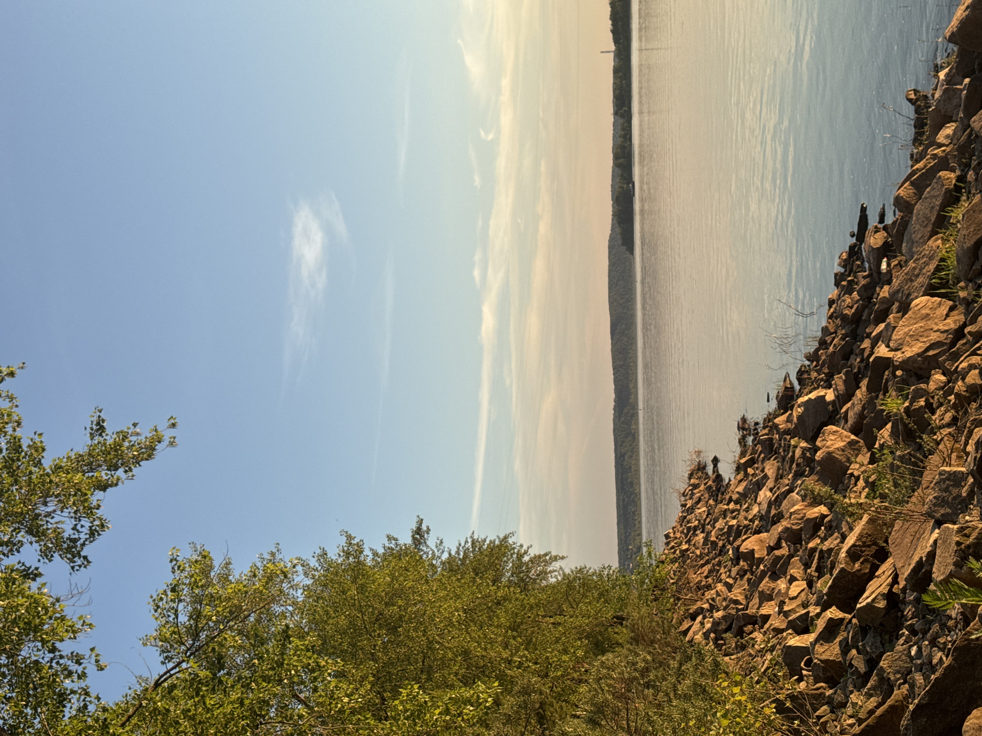 The image shows a tranquil waterscape with a rocky shore in the foreground. Lush green trees with dappled sunlight line the left edge of the frame, their branches extending towards the clear blue sky. The water is calm, reflecting the soft hues of the sunset, with a faint silhouette of a tree-covered landmass visible on the horizon. A thin, wispy cloud drifts across the sky. A small vessel, possibly a barge, can be seen on the water in the distance, near the far shore. The scene is serene and natural, with no people or man-made structures other than the distant vessel and a tall, slender industrial smokestack visible on the far right horizon. The location is described as Noviy, Russia, likely depicting a waterfront area along a river or large lake.