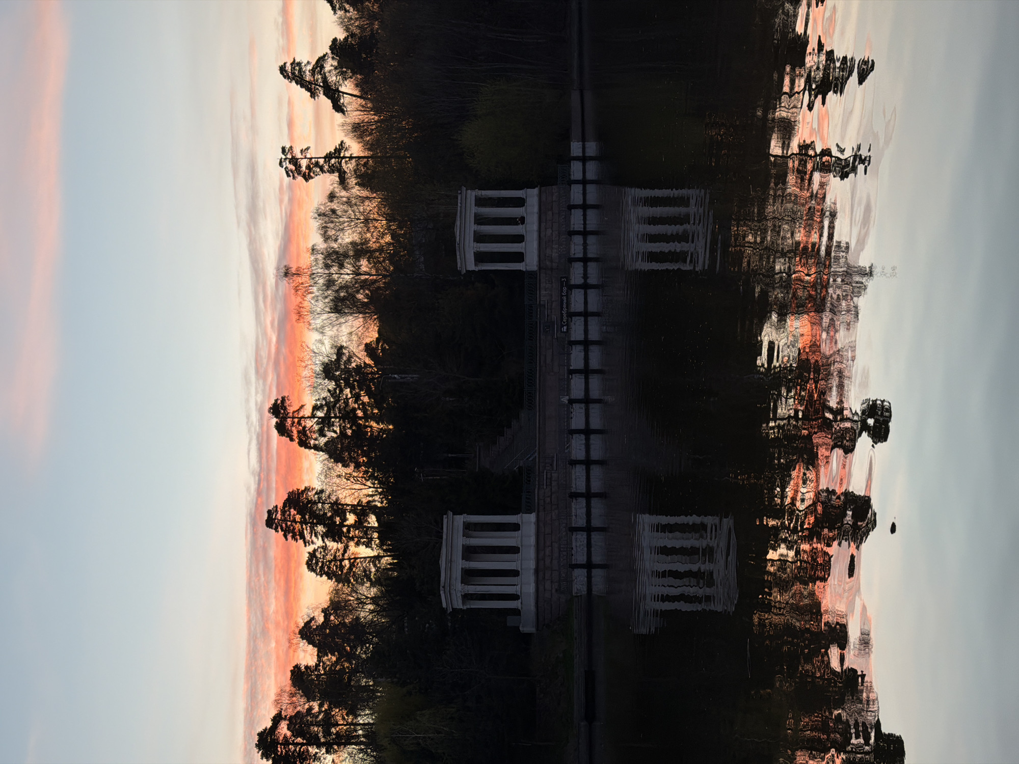 The image depicts a landscape scene likely captured during sunset or sunrise. The sky transitions from a pale blue to a soft orange-pink hue, suggesting either dawn or dusk. Silhouettes of trees are visible against the colorful sky.
In the middle of the image, there is a structure resembling a pavilion or a small building with architectural details. The presence of stairs suggests access to a higher level. The structure and the surrounding trees appear to be reflected in a body of water.
Given the reference to Khimki, Russia, and considering the structural features, the image likely shows the "Serebryany Bor" park in Khimki, with a view of the rotunda reflected in the water. There are no people visible in the image. The scene is static, capturing a moment of tranquility.